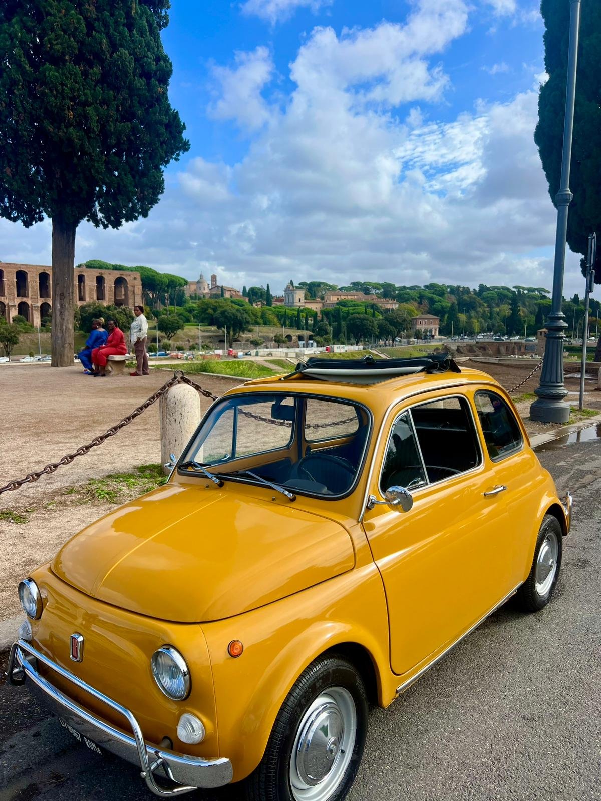 Auto d'epoca gialla parcheggiata su strada con albero e lampione, tre persone sedute su panchina in un parco, con colline e edifici storici sullo sfondo sotto un cielo nuvoloso.