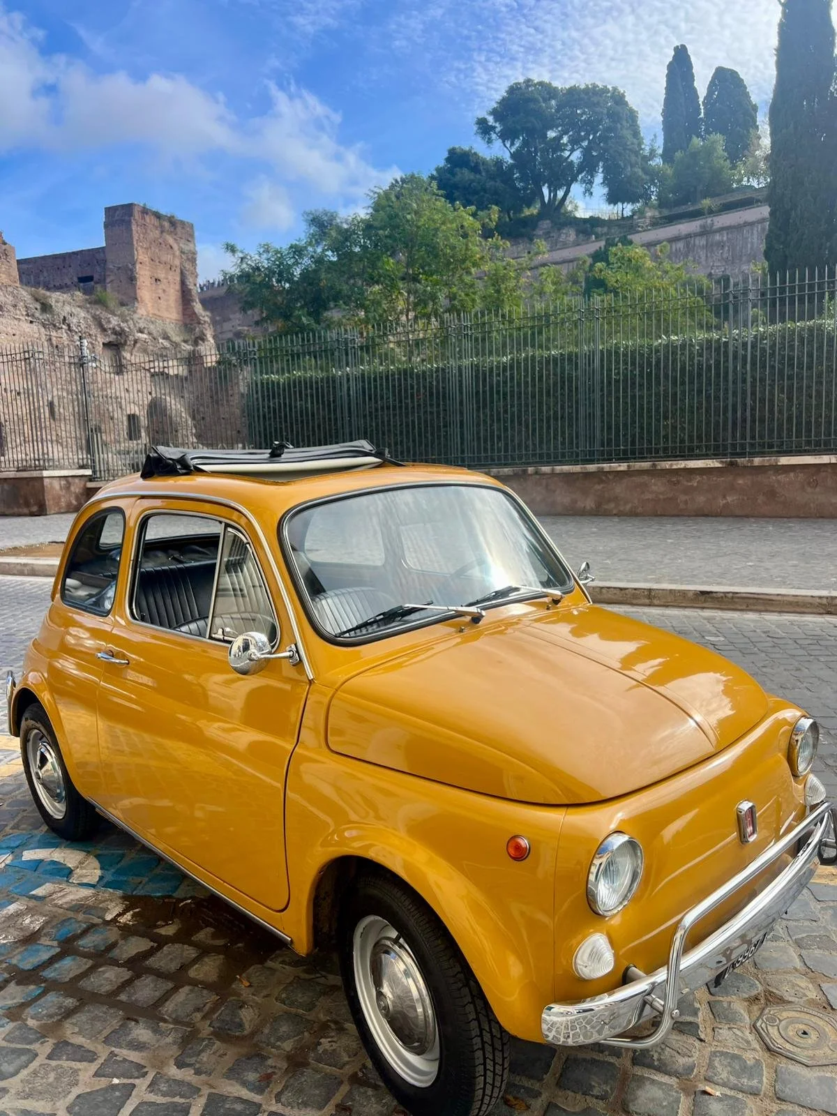 Auto d'epoca gialla parcheggiata su una strada di ciottoli con uno sfondo di pareti e una collina con alberi e resti di un castello o rovina storica sotto un cielo blu con nuvole