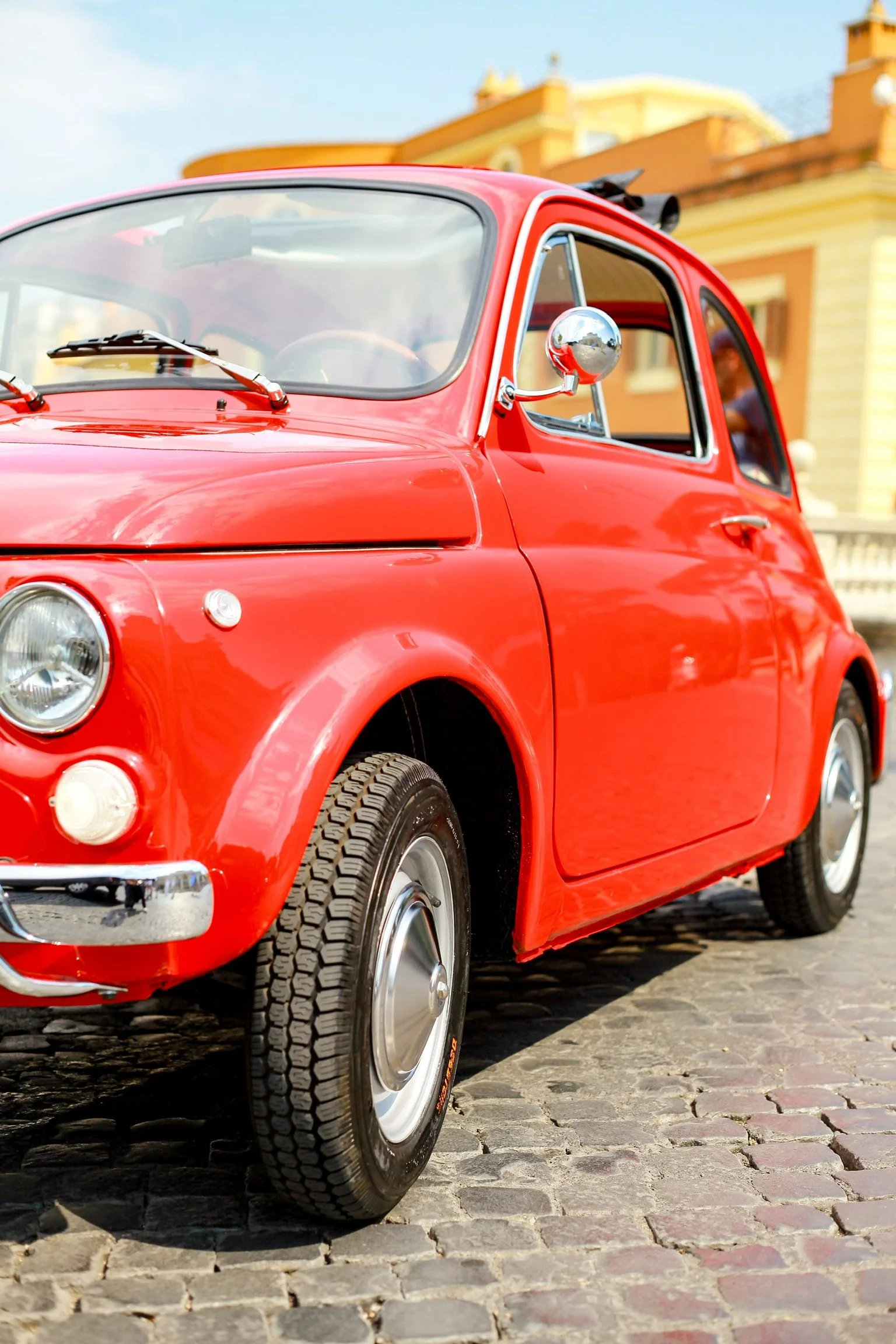 Auto rossa d'epoca parcheggiata su una strada in pietra con edifici storici sullo sfondo.