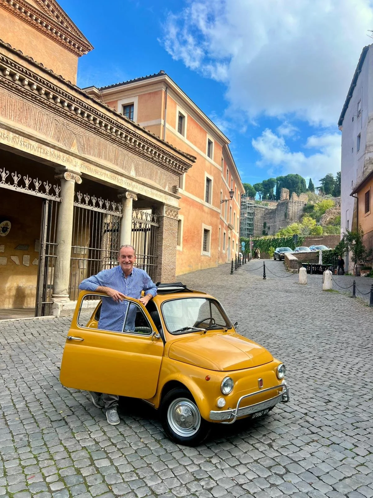 Un uomo felice con una camicia a quadri azzurra si appoggia a una piccola auto gialla parcheggiata su una strada acciottolata nel centro storico di una città italiana con edifici colorati e un cielo azzurro con nuvole sparse.