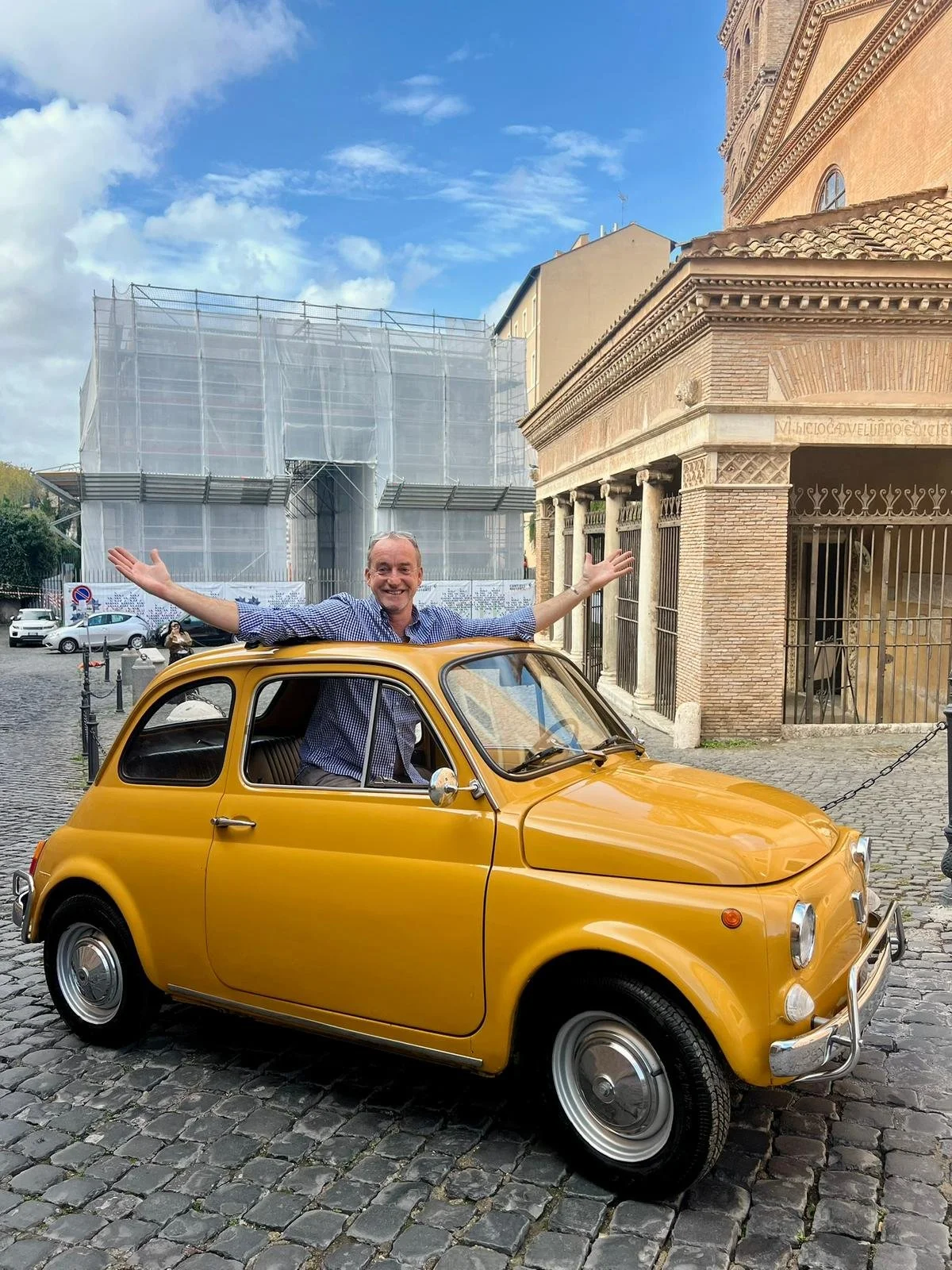 Uomo felice che si trova dentro una piccola auto gialla vintage, con le braccia aperte, davanti a edifici storici e un cielo blu.