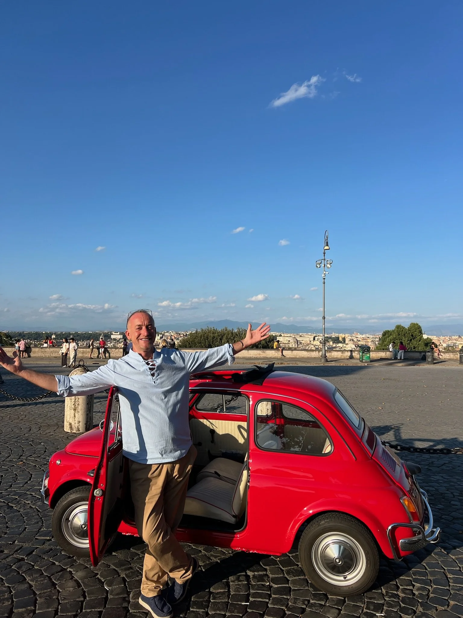 Uomo sorridente con braccia aperte davanti a una piccola auto rossa, con una città in lontananza e un cielo sereno.