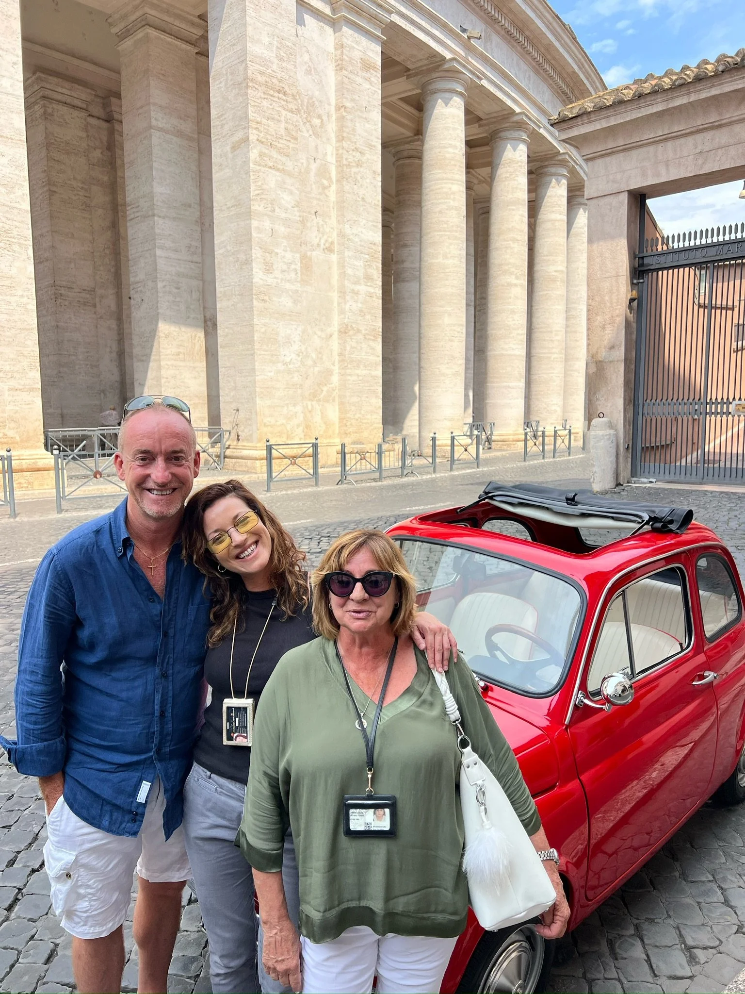 Tre persone sorridenti davanti a una piccola auto rossa d'epoca, con un edificio storico con grandi colonne sullo sfondo.