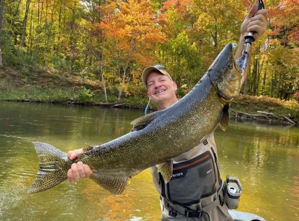 A man with a cap smiling while holding a large fish on a fishing line in a river surrounded by colorful autumn trees.