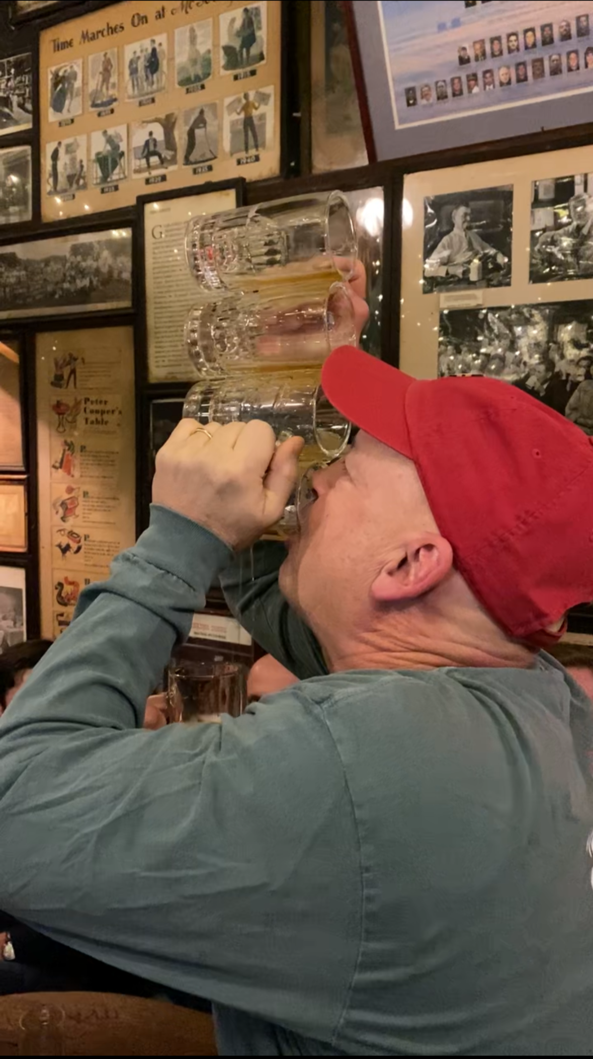 A man wearing a red cap and gray shirt is holding five beer glasses stacked on top of each other to his mouth in a bar, surrounded by vintage photographs and posters on the wall.