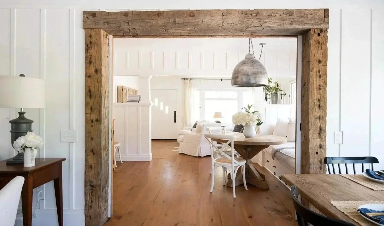 Interior view of a bright, cozy living and dining area with white walls, wooden floors, and rustic wood beam framing the entrance to the living room. The space features a round wooden dining table with white and black chairs, a gray pendant light, an