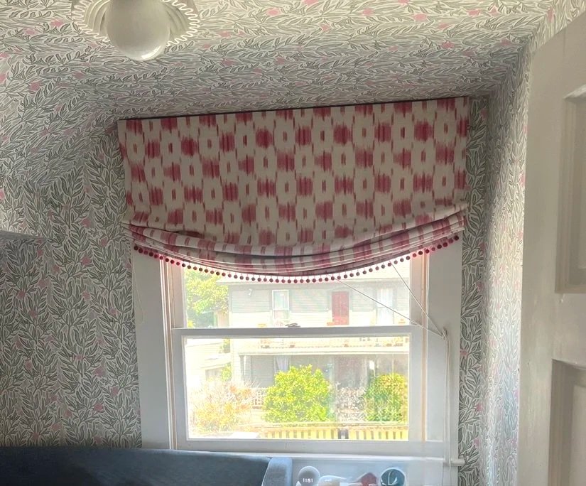 A window with a red and white patterned valance and a matching curtain, overlooking a colorful residential neighborhood with trees.