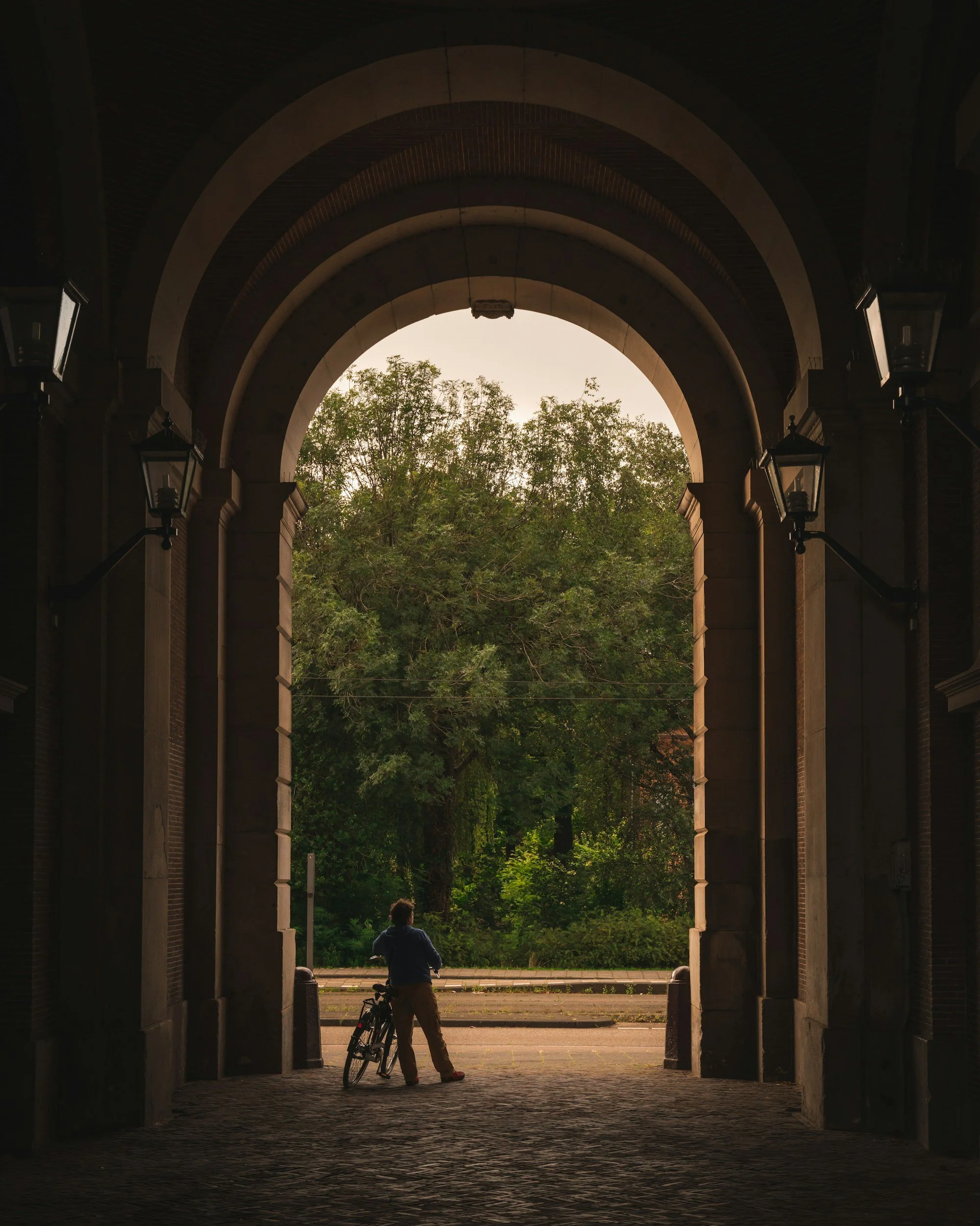 A person standing with a bicycle under an arched brick passageway, looking out at a tree-lined street during sunset.