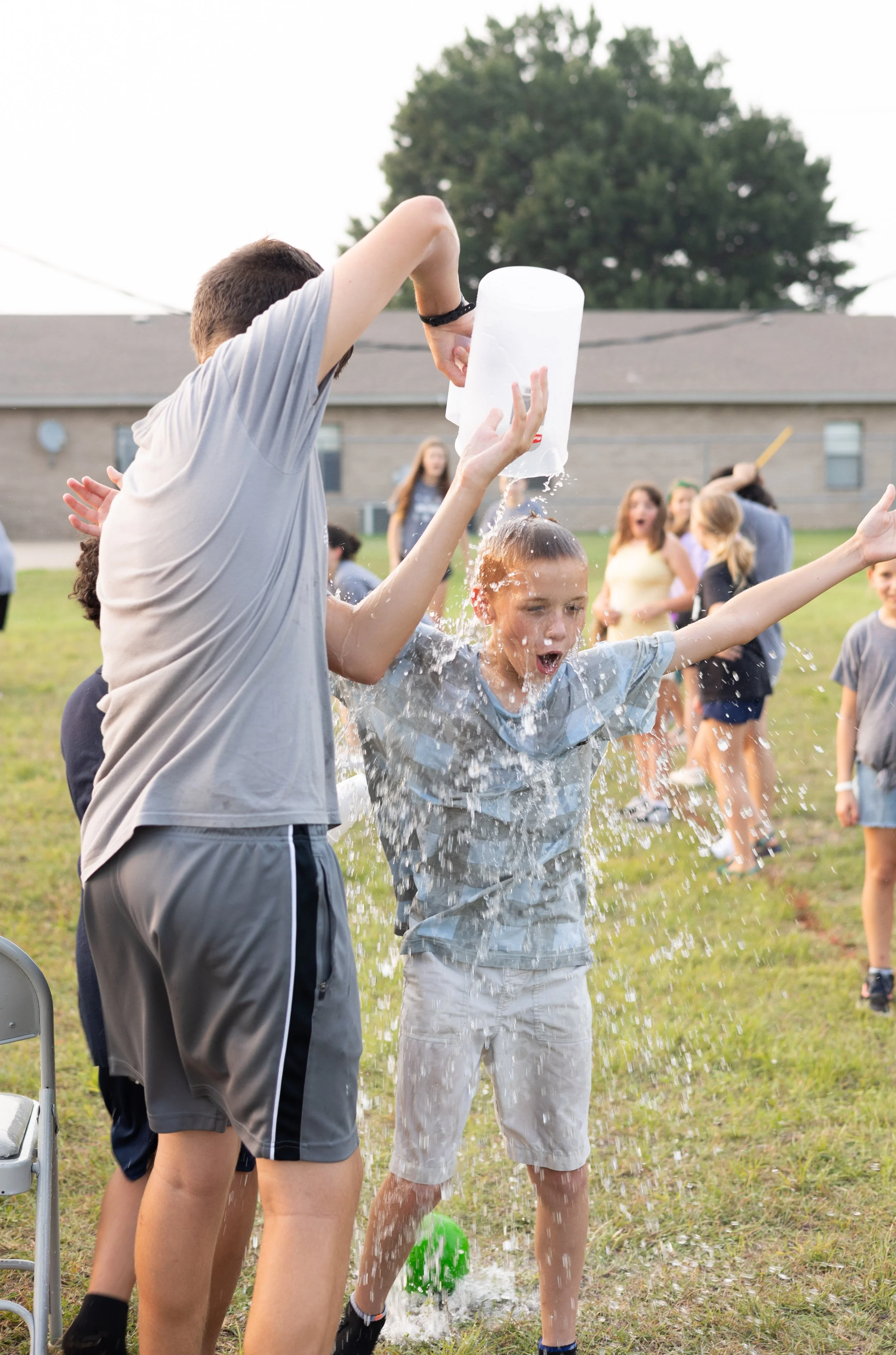 Two boys are standing outside, pouring water on each other during a summer activity. Other children are visible in the background, and the scene takes place on a grassy field with a building and a tree in the distance.