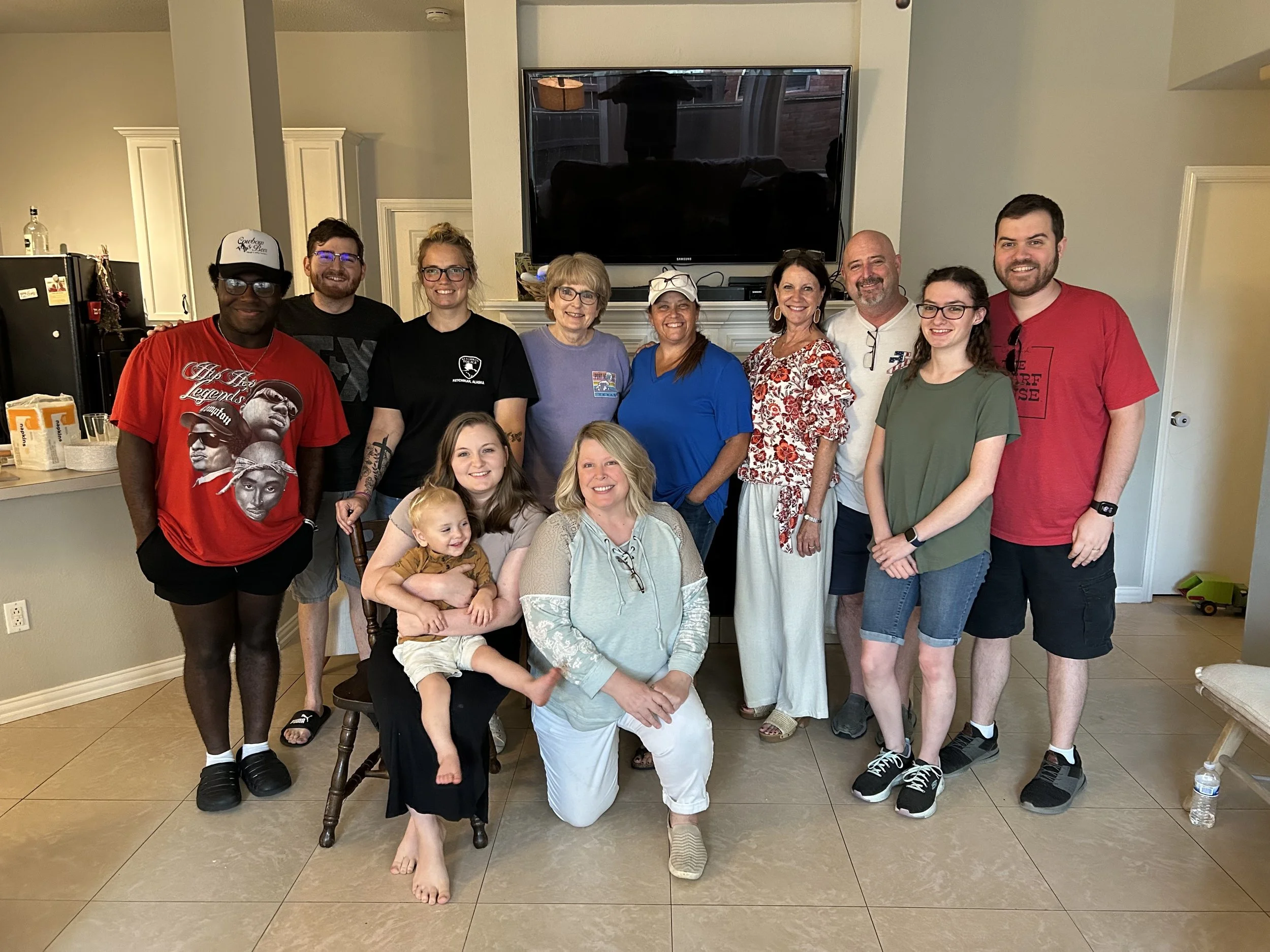 Group of twelve people, including adults and a child, gathered indoors in front of a fireplace and TV, smiling for the photo.