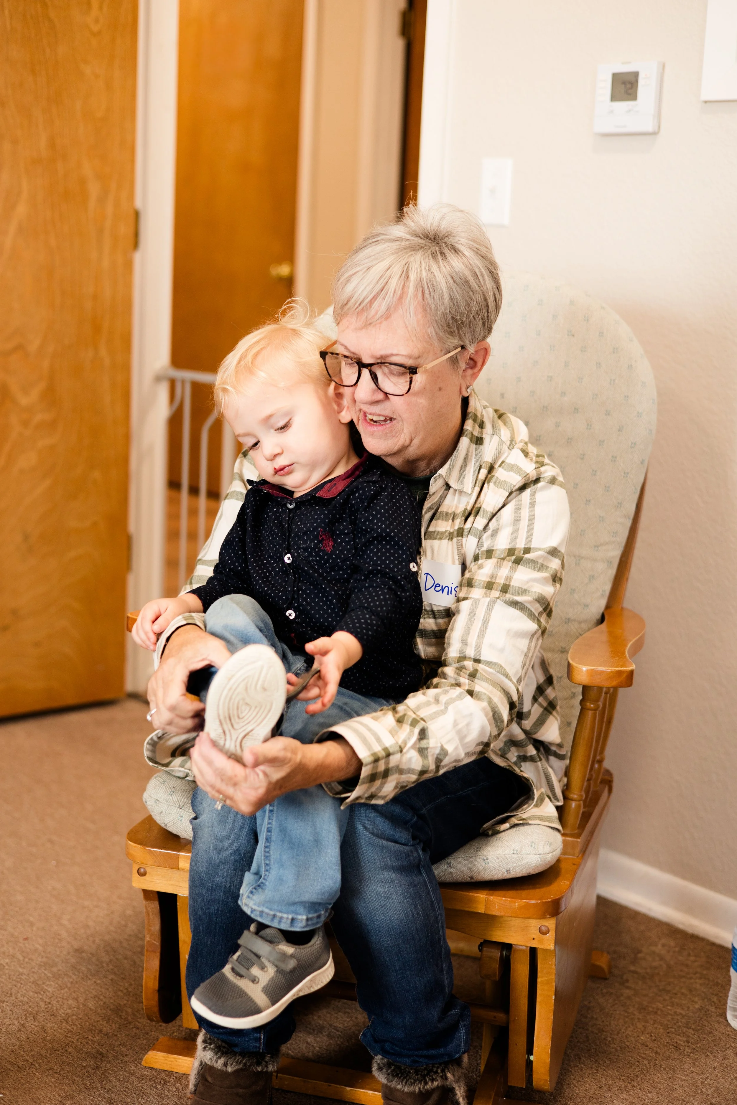An elderly woman with glasses and a plaid shirt is sitting on a wooden chair, holding a young blonde boy on her lap who is trying on a shoe. The woman looks slightly scrunched up as she helps the boy.