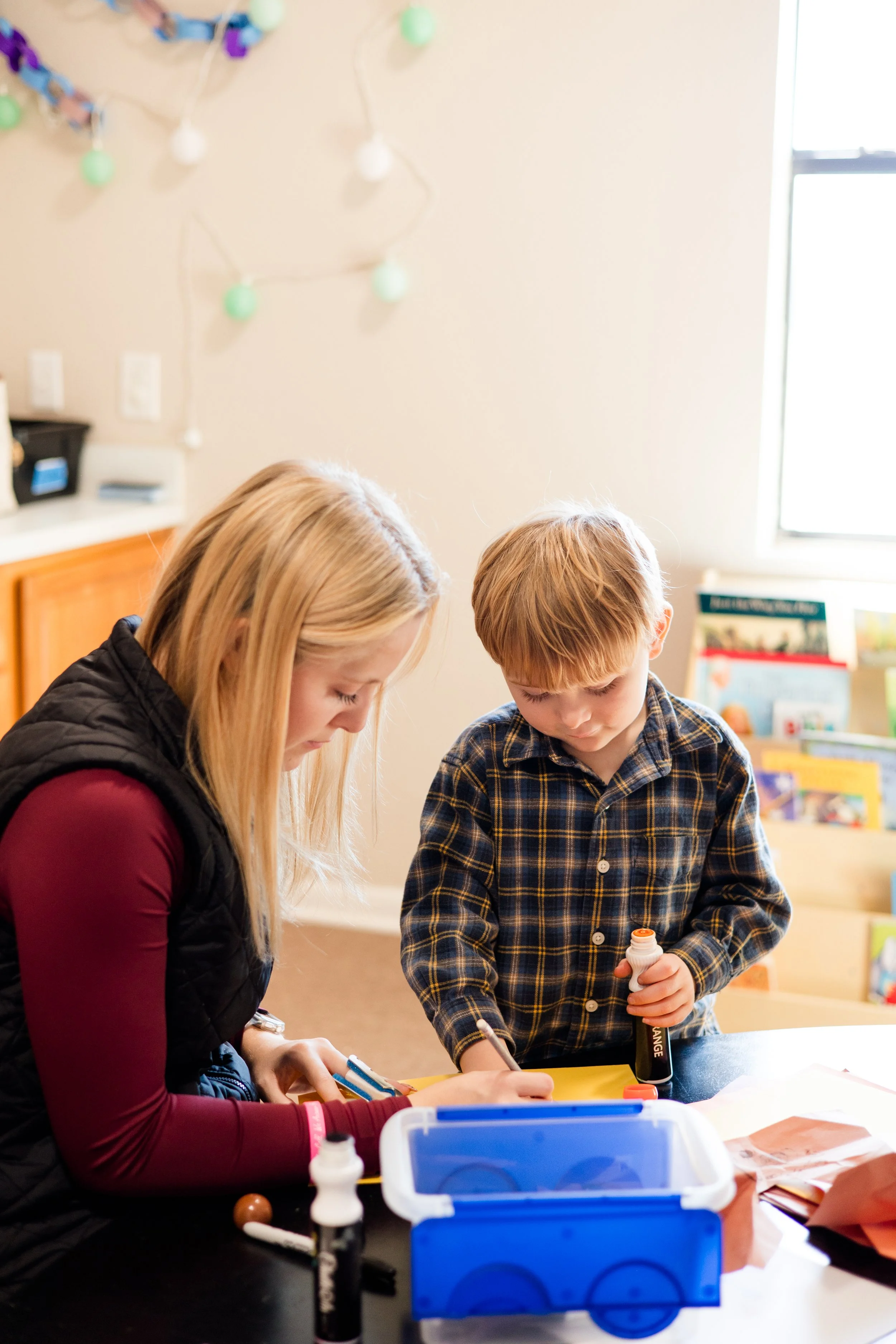 A woman and a young boy working together at a table, with various craft supplies, in a room decorated with string lights and books