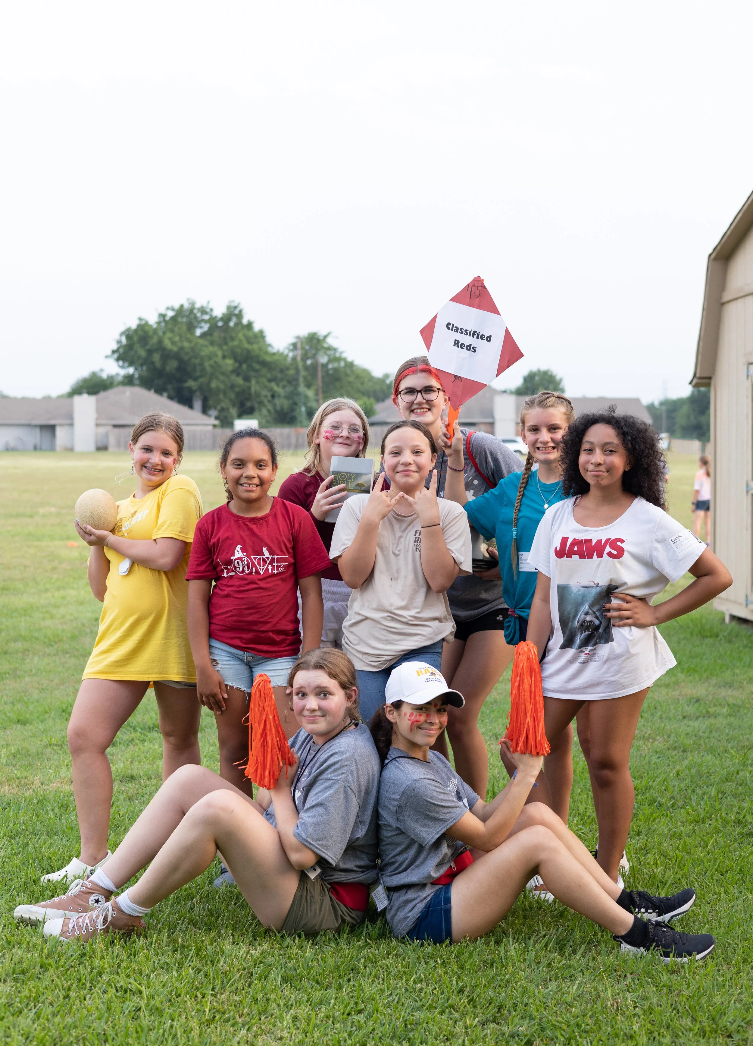 Group of children and teenagers outdoors, some smiling, holding a sign that reads 'Classified Reds' and a pumpkin, with some wearing face paint and holding pom-poms, on a grassy field with houses and trees in the background.