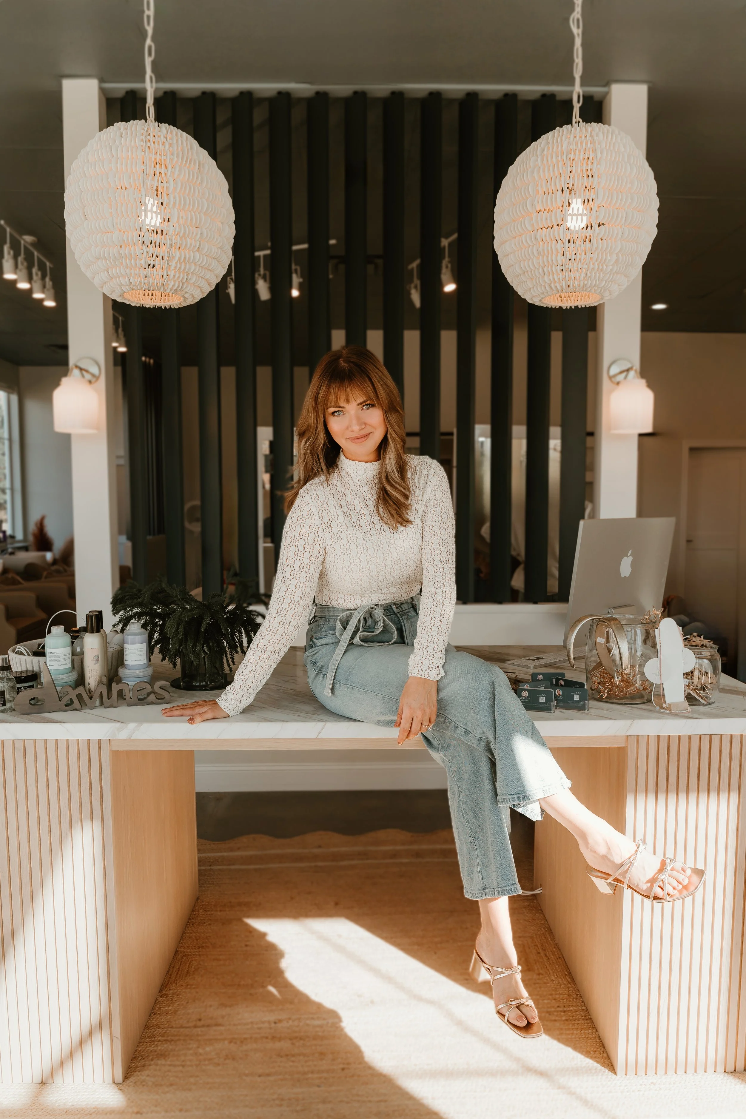 Crystal Scott, founder of Adrift Hair Co. in Wake Forest. Balayage and Great Lengths Extensions certified salon. Woman sitting on a reception desk in a modern, well-lit room with hanging pendant lights and black vertical slats on the wall behind her.