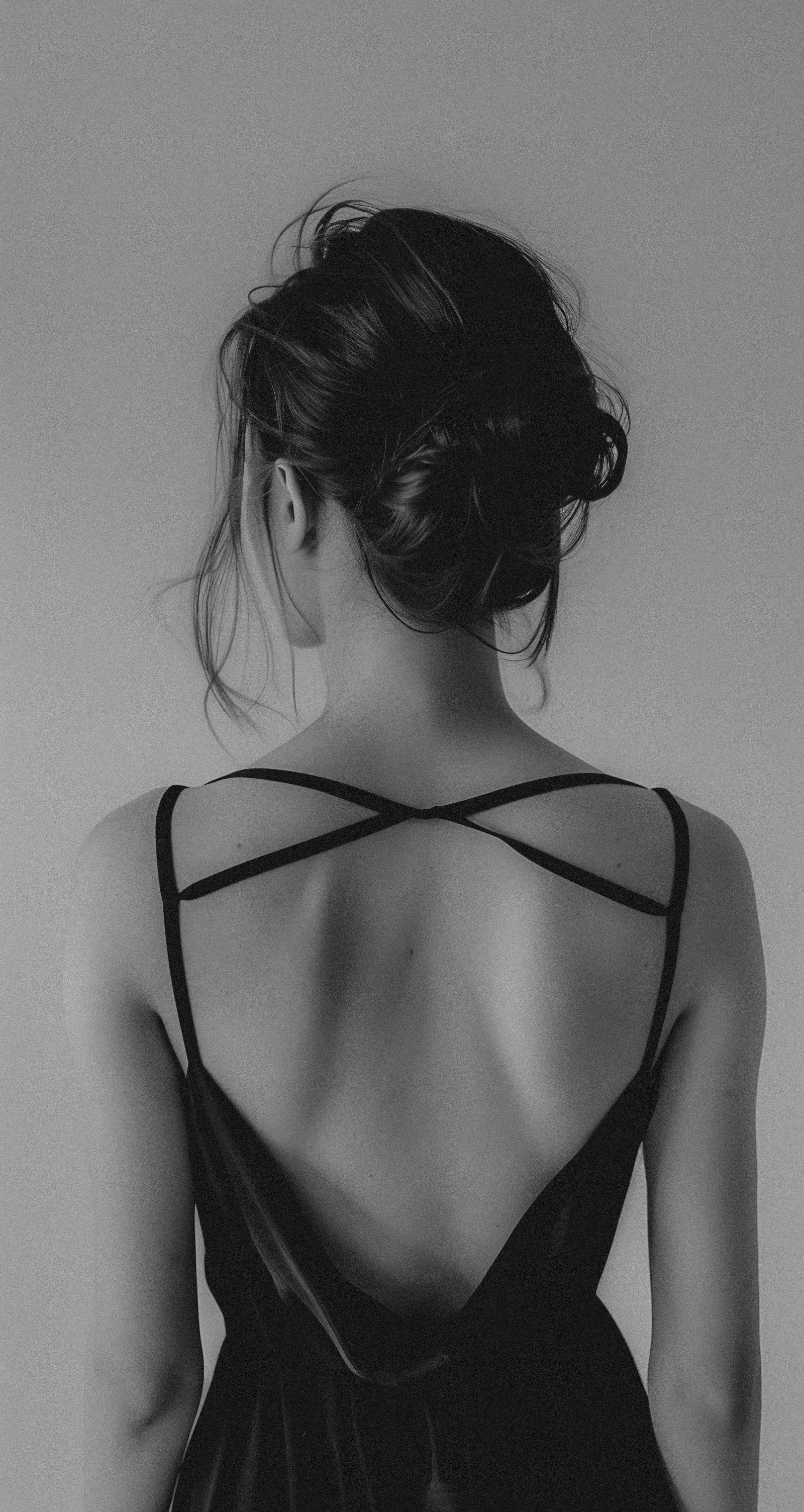 A woman with dark hair styled in an updo, viewed from the back, wearing a dress with thin crisscross straps on her shoulders, in black and white.