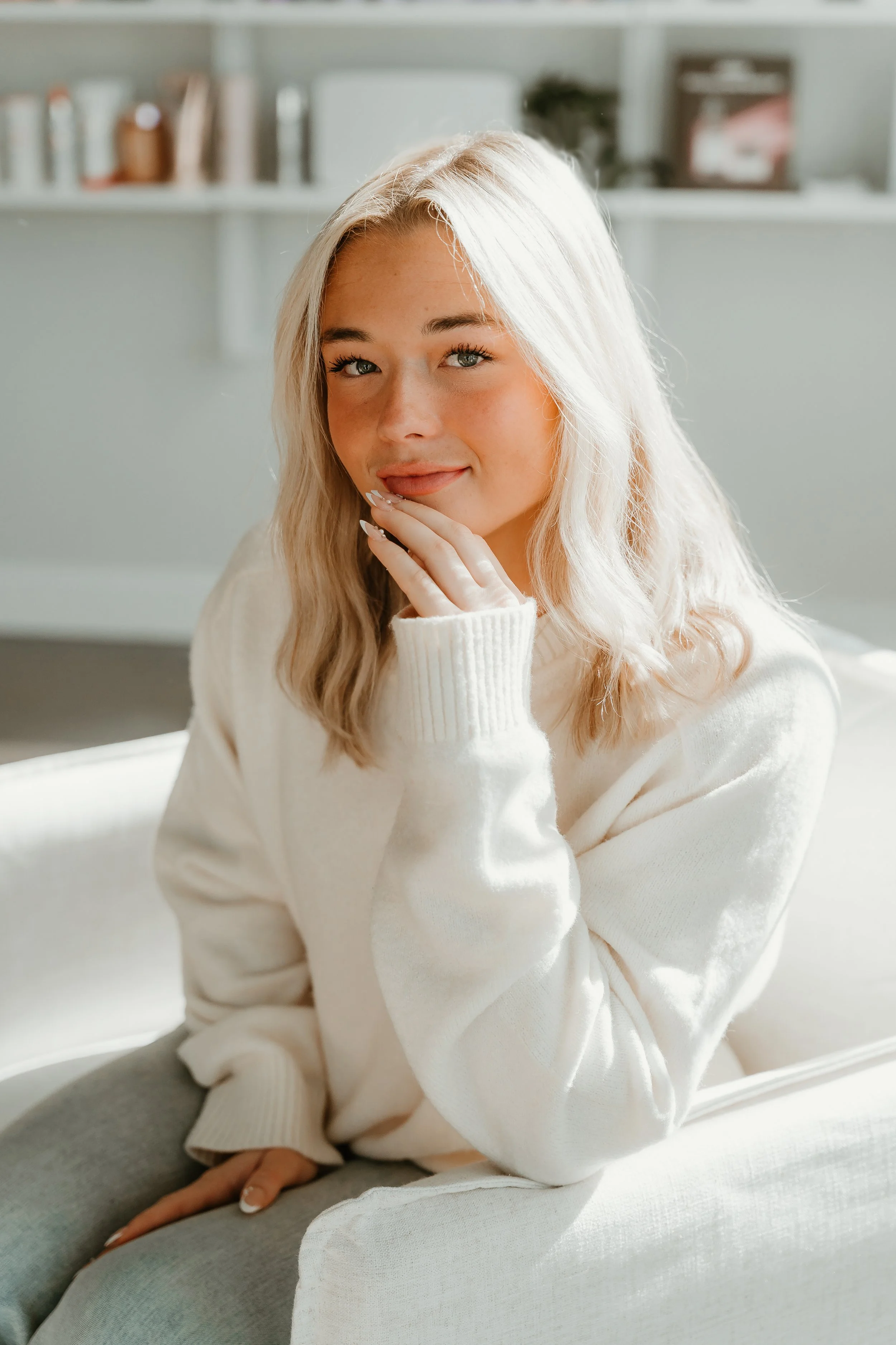 A young woman with blonde hair, wearing a cream-colored sweater, sitting on a white chair in a well-lit room with a bookshelf in the background.