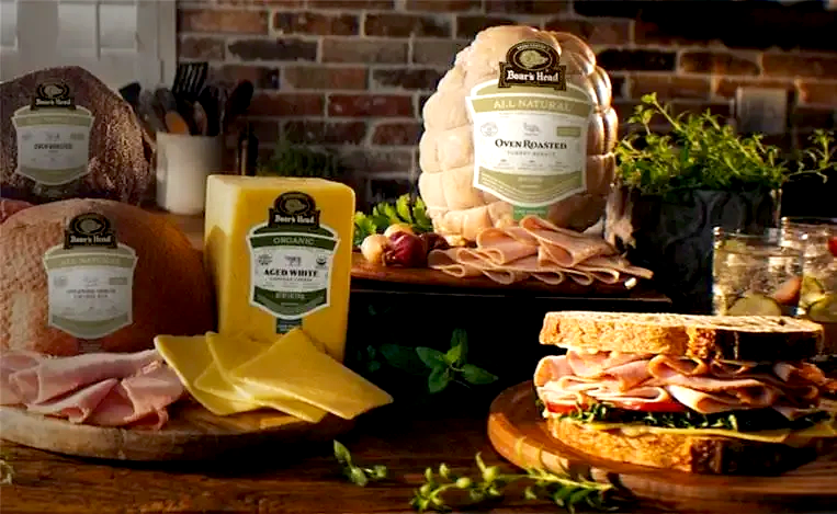 Variety of deli meats and cheeses on wooden boards with a cooked whole chicken and a brick wall background