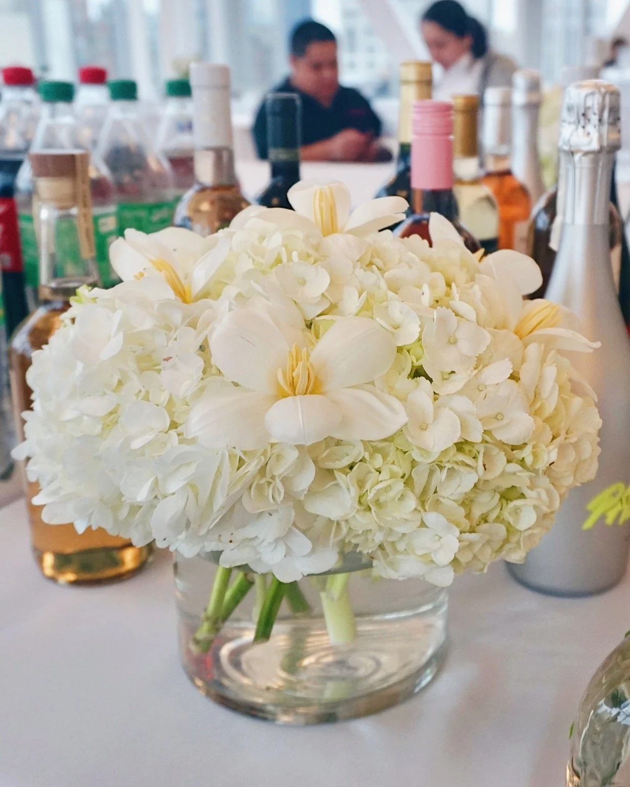 White hydrangea and orchid flower bouquet in a clear glass vase on table with bottles in the background