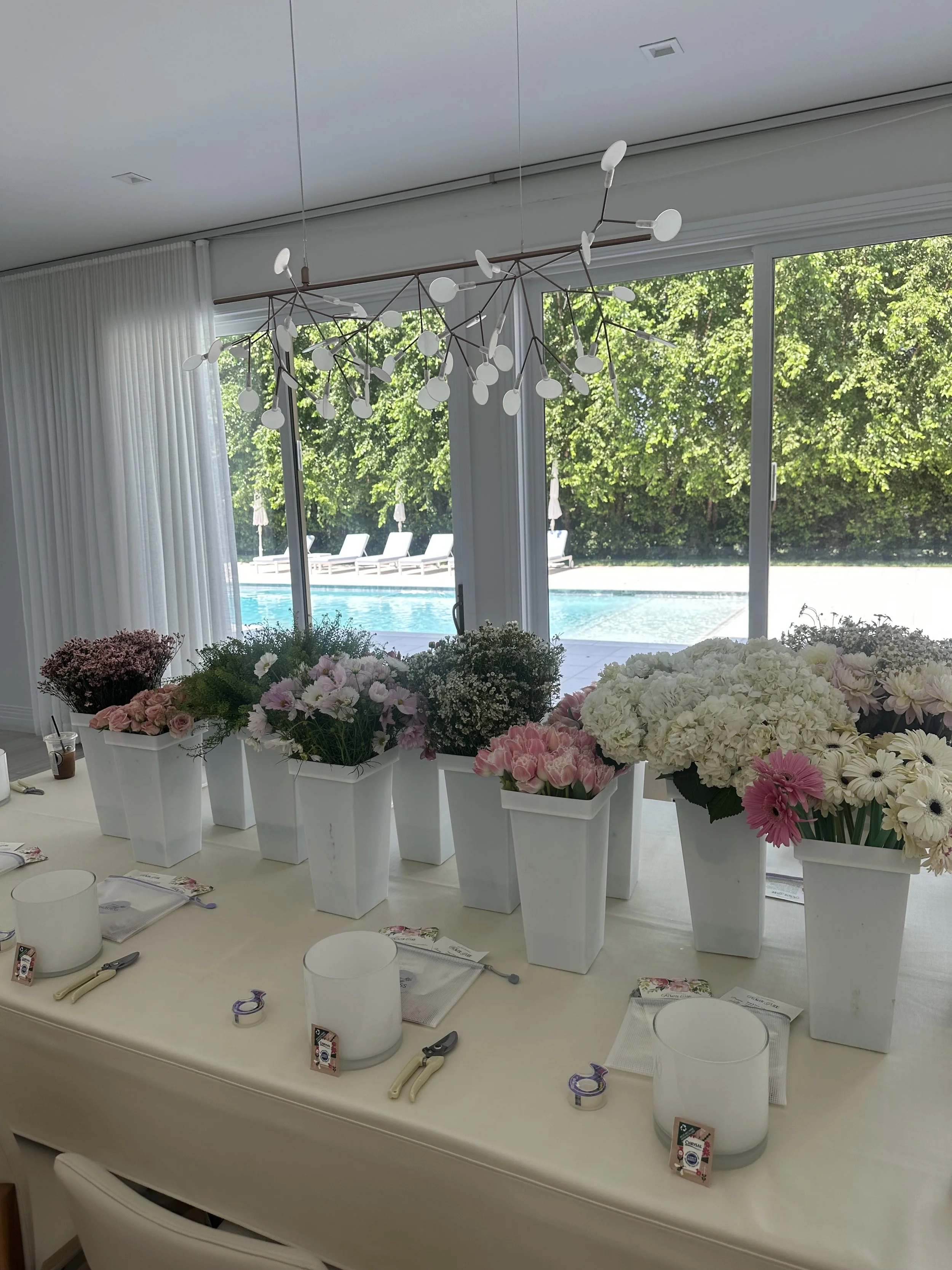 A dining table decorated with seven white vases filled with pink and white flowers, set in front of large glass sliding doors overlooking a pool and greenery outside, with a modern chandelier hanging from the ceiling.
