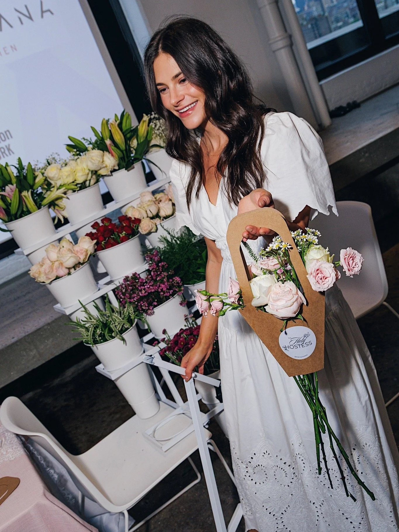 Woman in white dress holding a bouquet of pink and white flowers at a flower stand.