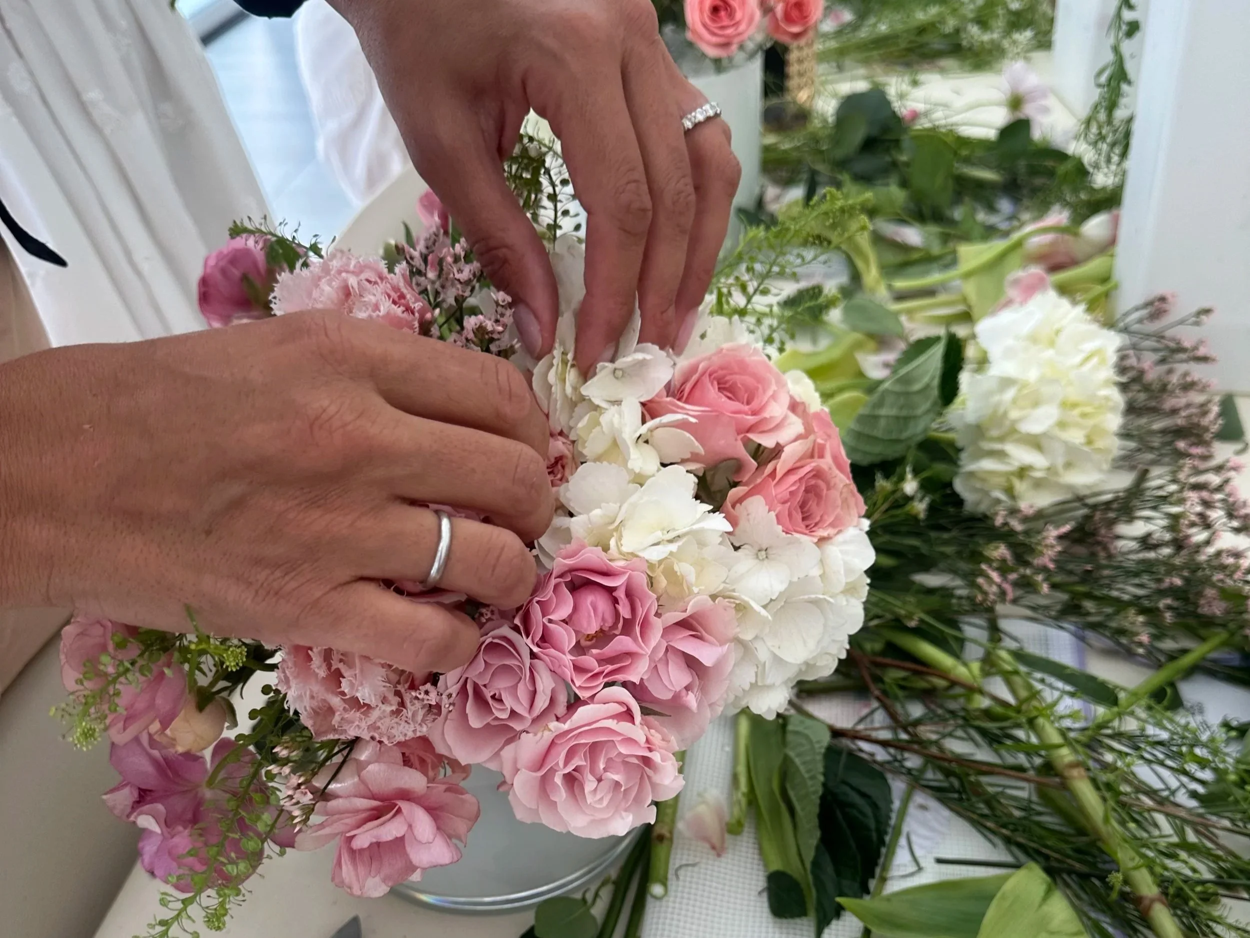Person arranging pink, white, and lavender flowers in a bouquet with greenery, on a table covered with flower stems and leaves.