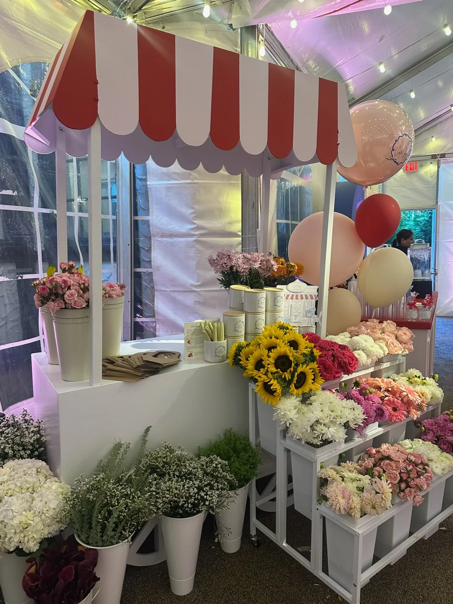 A flower stall under a canopy with a red and white striped awning, displaying various freshflowers including sunflowers, roses, and daisies, with balloons in the background at an event.