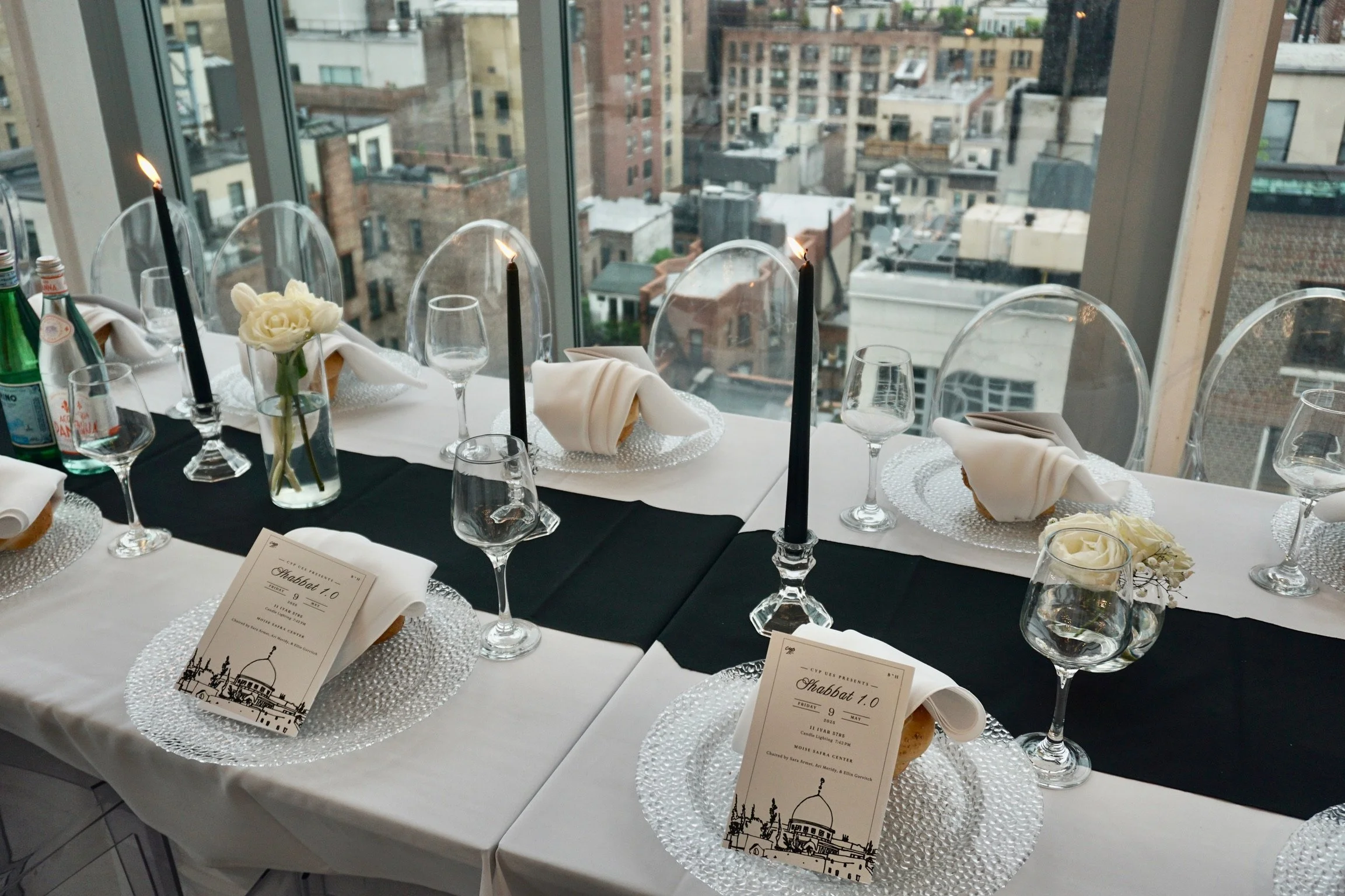 A elegantly set dining table with white tablecloths, black and white napkins, clear glass plates, wine glasses, and black candles in candleholders, decorated with white flowers, in a high-rise restaurant with city view windows.