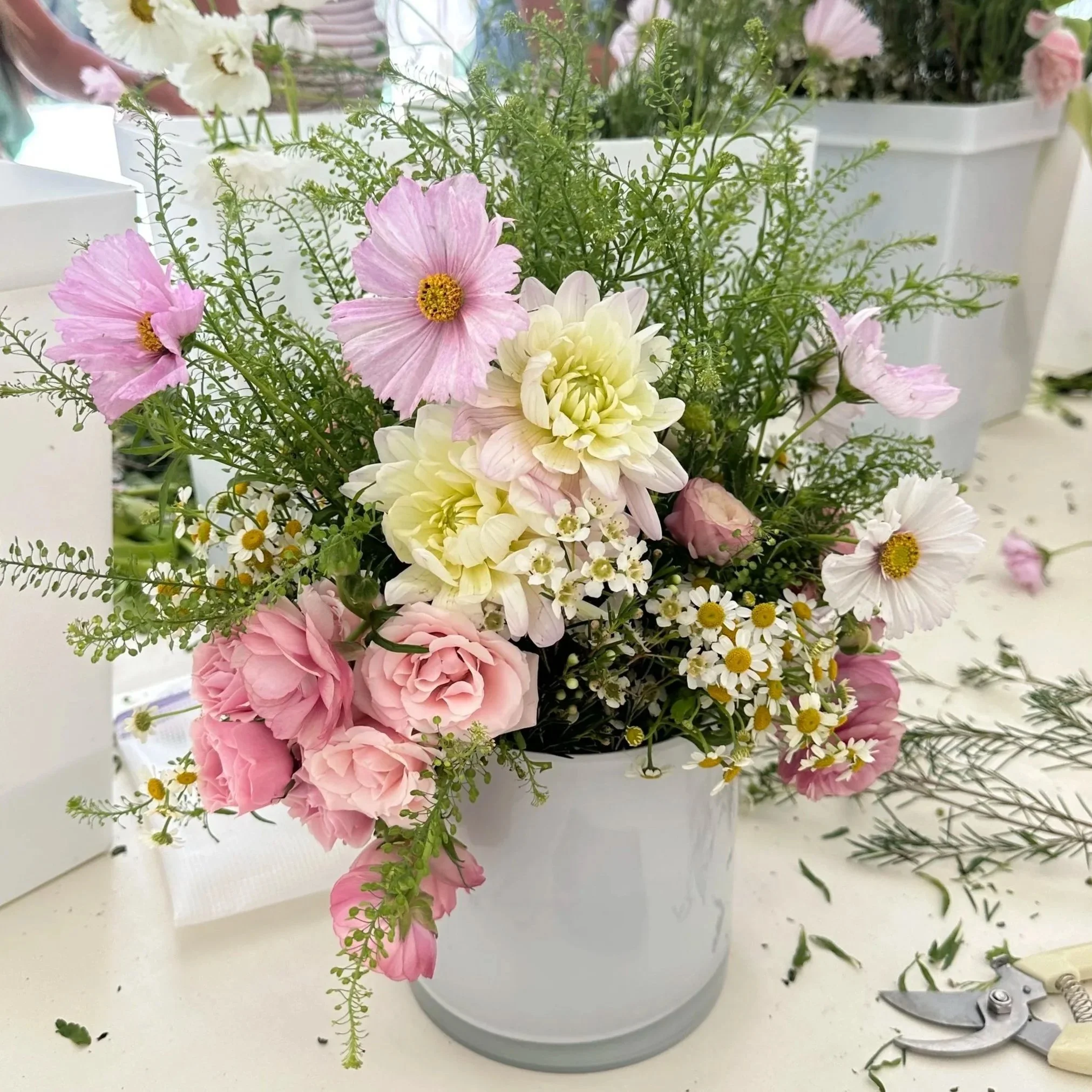 A white vase filled with pink, white, and cream flowers, including daisies, dahlias, and roses, along with green foliage.