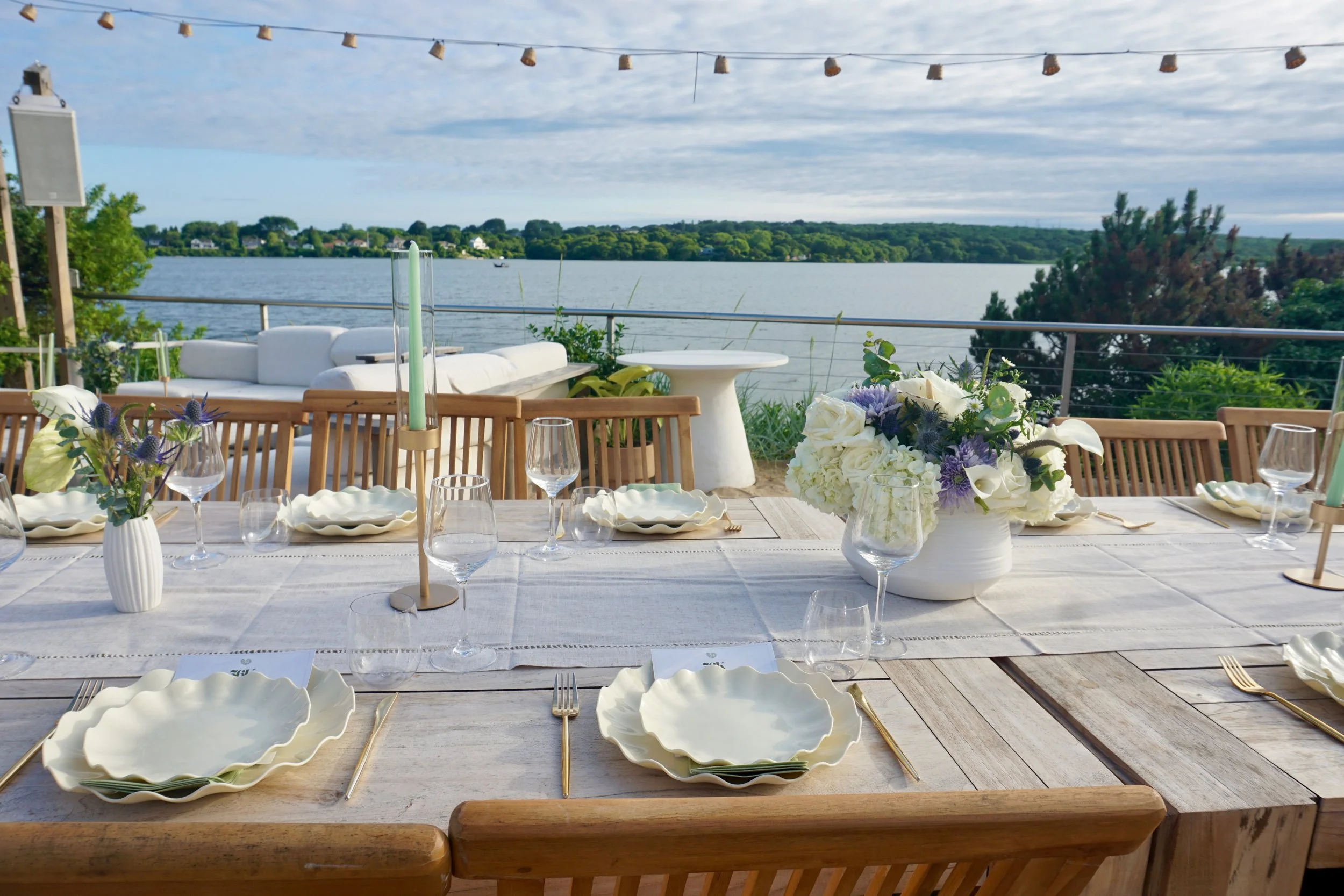 Outdoor waterfront dining setup with a long wooden table, white ceramic plates, gold cutlery, glassware, floral centerpieces, and candles, overlooking a lake with trees and houses in the background under a partly cloudy sky.