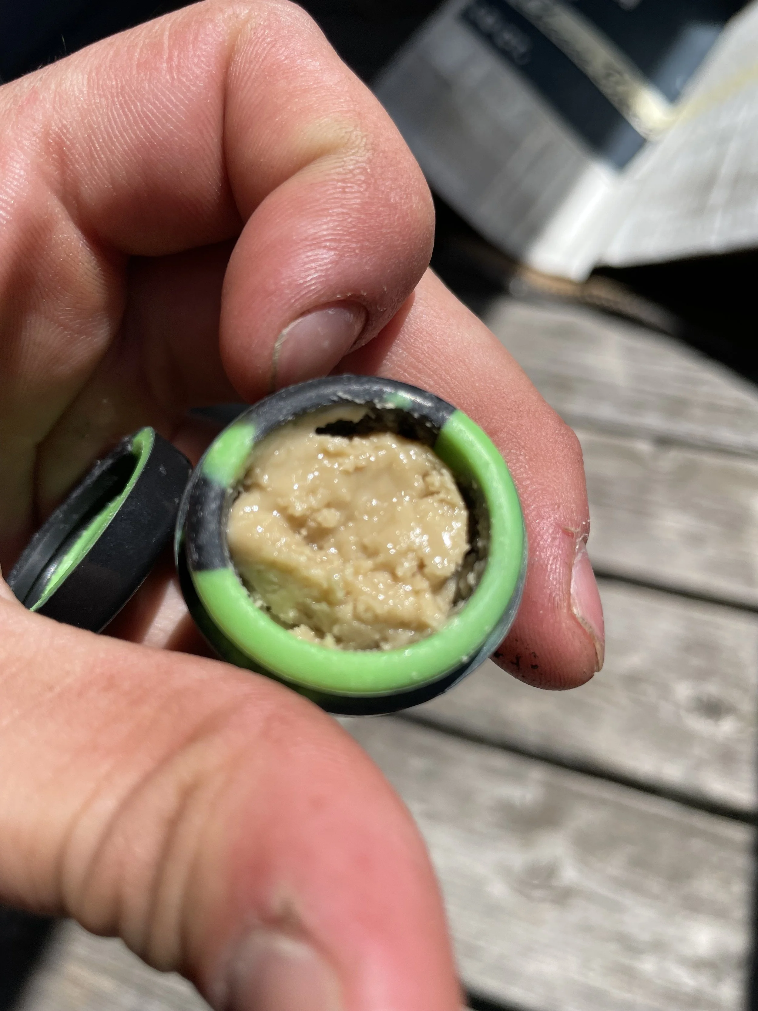 Close-up of a person's hand holding a small container with beige, sticky substance inside and green accents on the container.