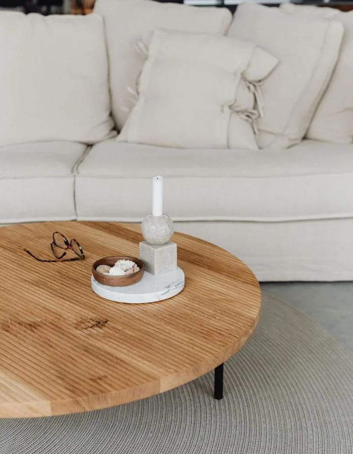 A wooden coffee table with a pair of glasses, a small bowl of decorative objects, and a marble candle holder with a candle inside. In the background, a white sofa with multiple white cushions.