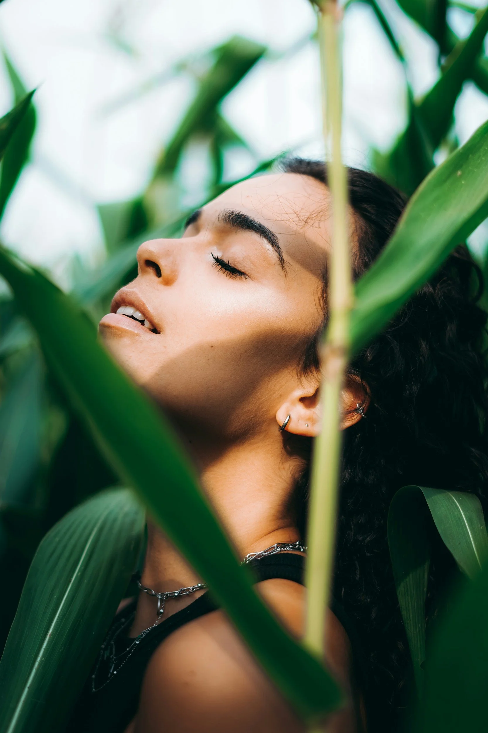 A woman with dark, curly hair and pierced ears, wearing layered necklaces and a black top, with her eyes closed, surrounded by tall green plants.