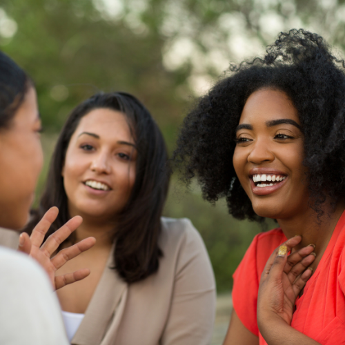 Three women are outdoors, engaged in conversation. One woman with natural curly hair is smiling and touching her chest, another woman with straight dark hair is gesturing with her hand, and the third woman's face is partially visible.