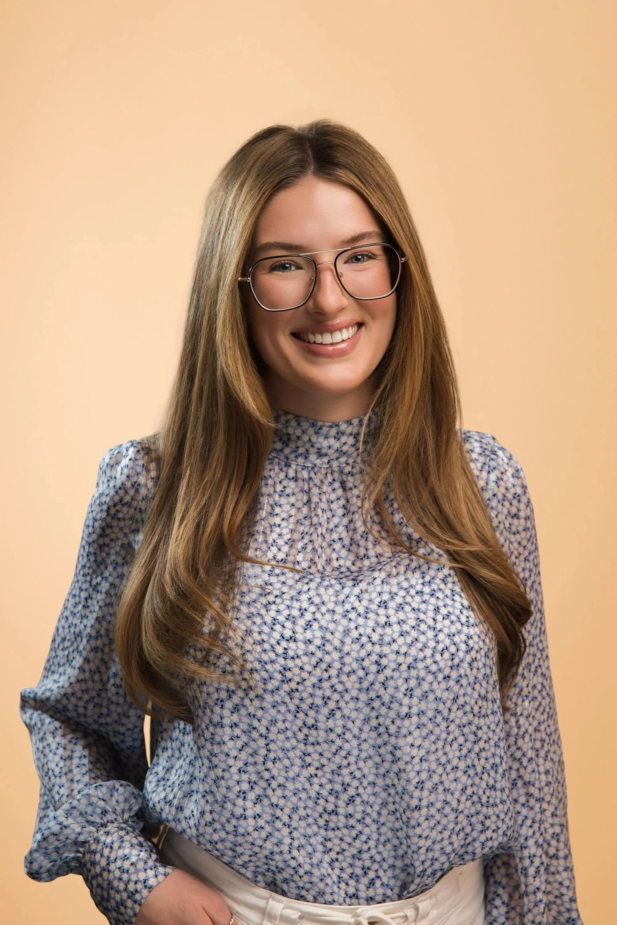 A young woman with long brown hair, wearing glasses, a floral blouse, and white pants, smiling against a beige background.