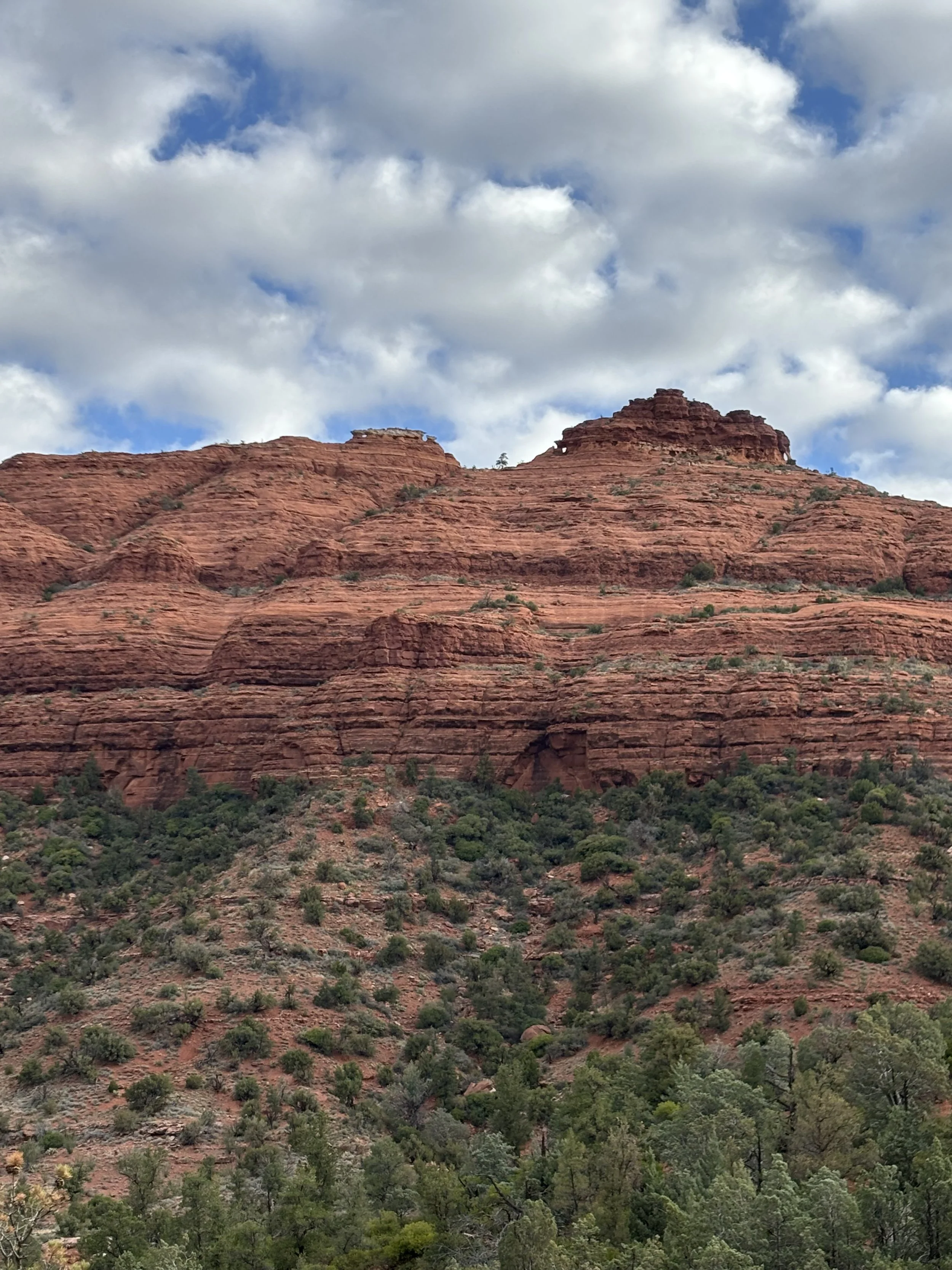 Red rock formations with green trees at the base under a partly cloudy sky.