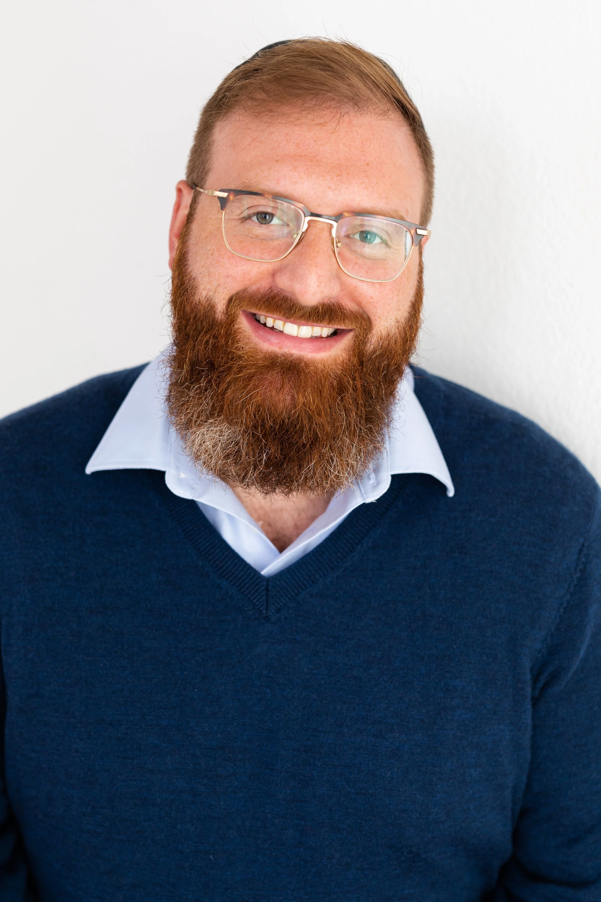 A man with red hair and a beard wearing glasses, a white collared shirt, and a navy blue sweater smiling against a white background.