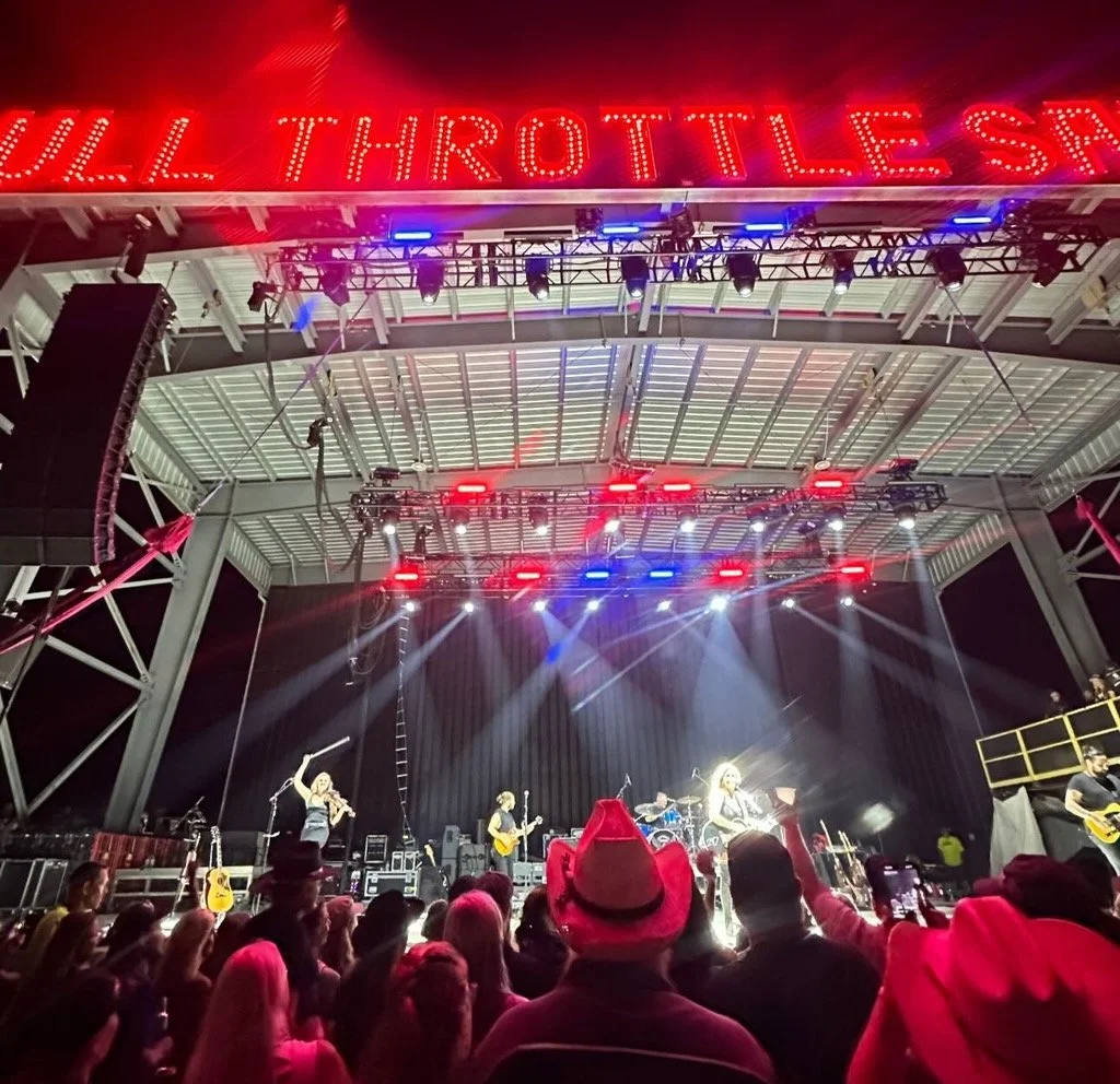 Concert stage with performers and audience at night, illuminated by stage lights and neon sign reading 'VILL THROTTLE' in the background.