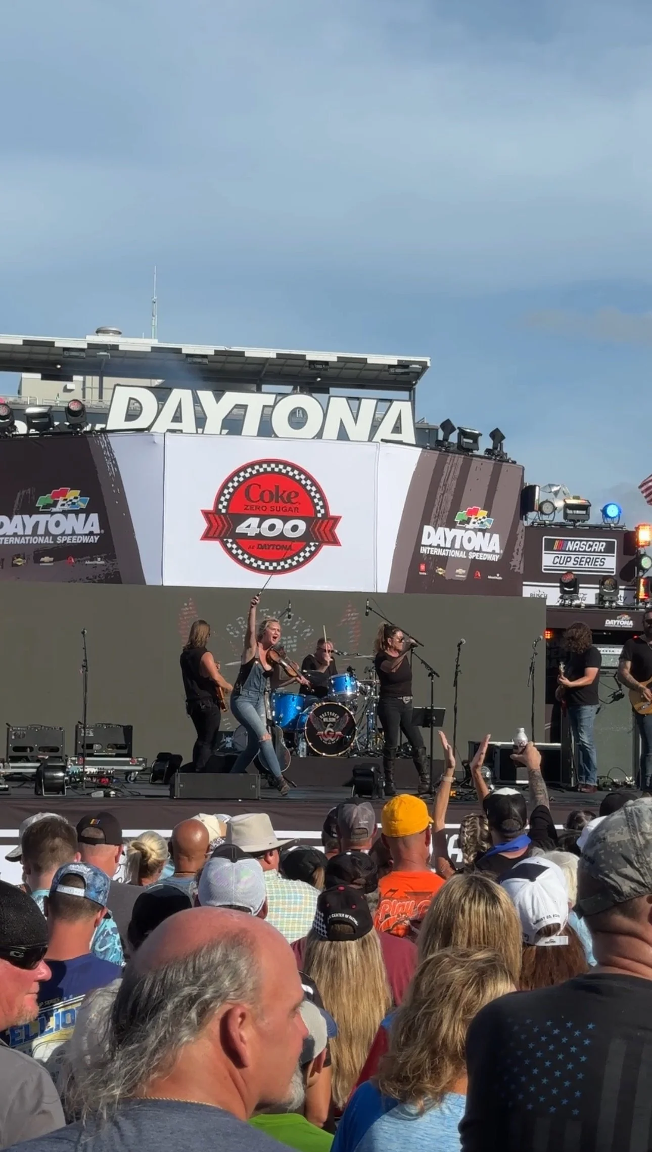 Live concert at Daytona International Speedway during a NASCAR event with a band performing on stage, crowd in attendance, and race-related signage in the background.
