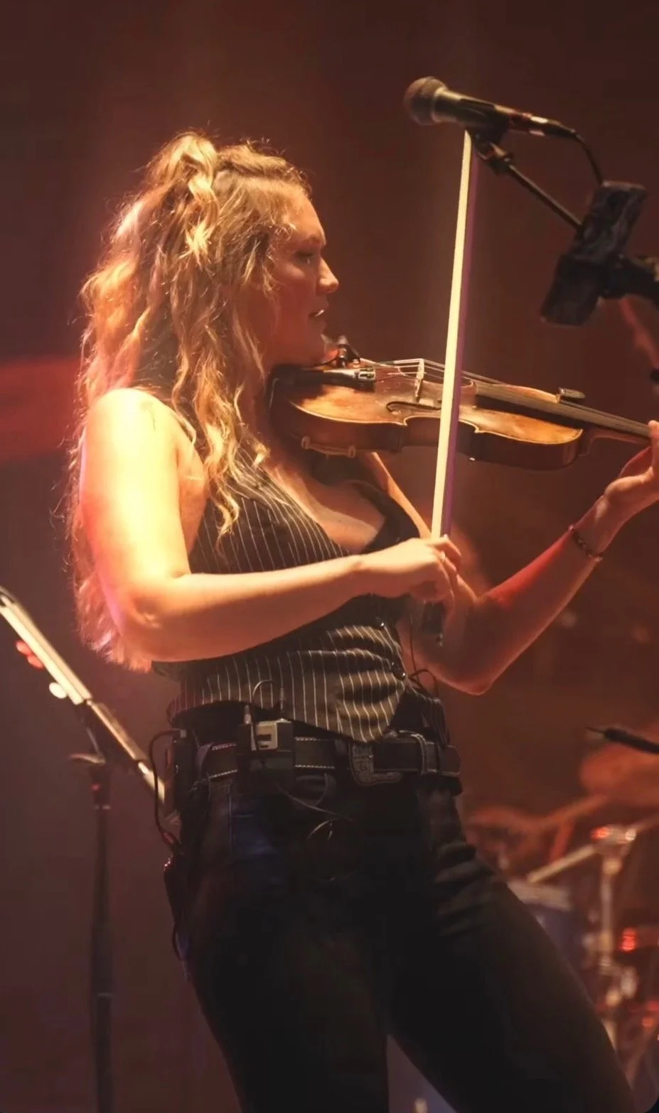 A woman playing the violin on stage during a concert, illuminated by red stage lights.