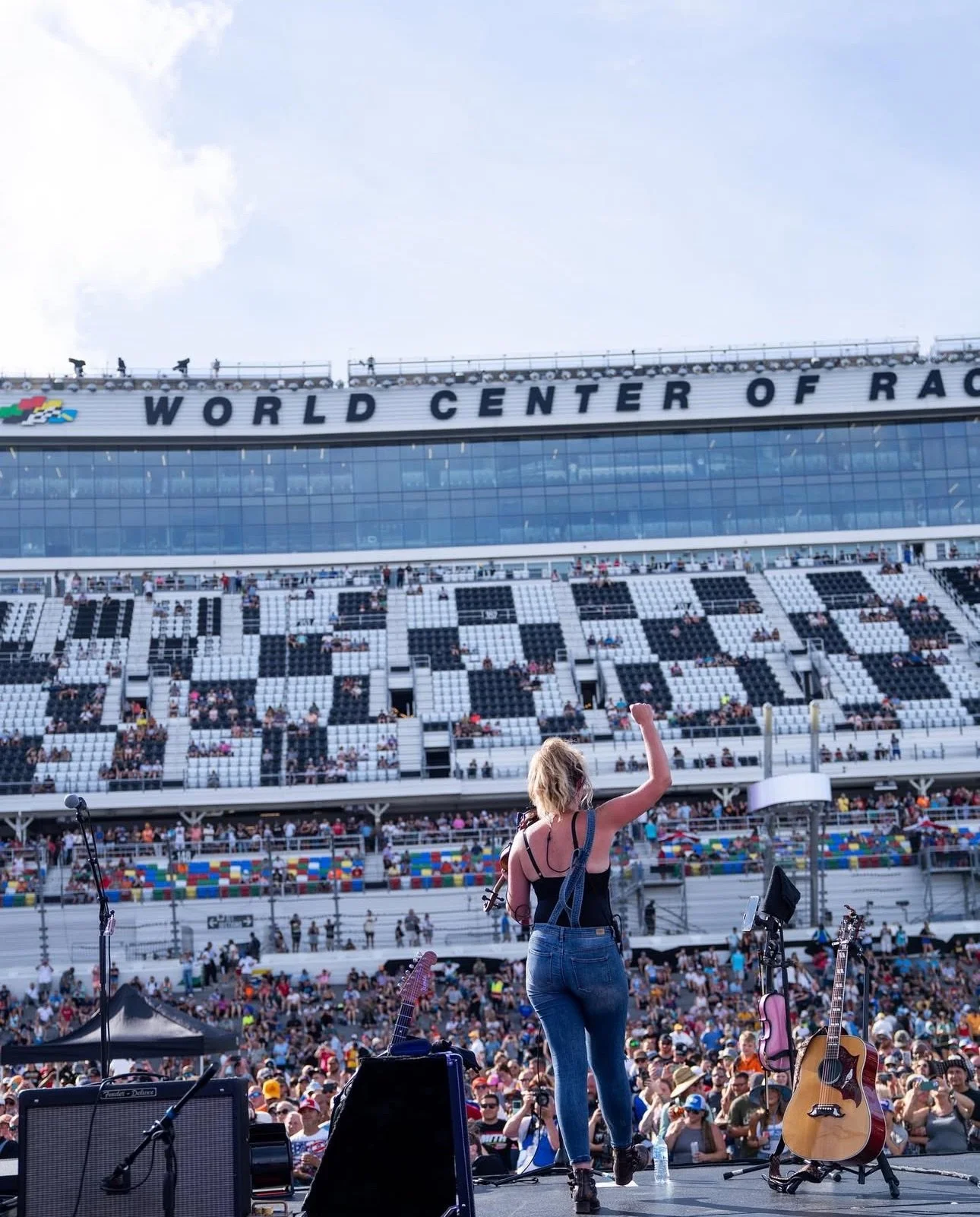 A female performer on stage at a large race track stadium, preparing to sing with guitars and microphones, during a concert at the World Center of Racing.