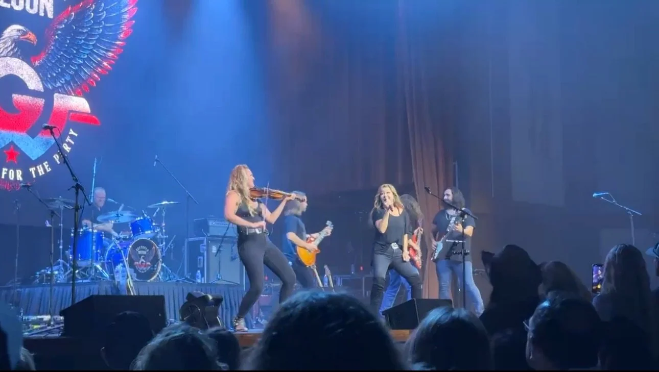 Live music concert with a band on stage, including a female singer, violinist, guitarist, and drummer, performing in front of an audience.