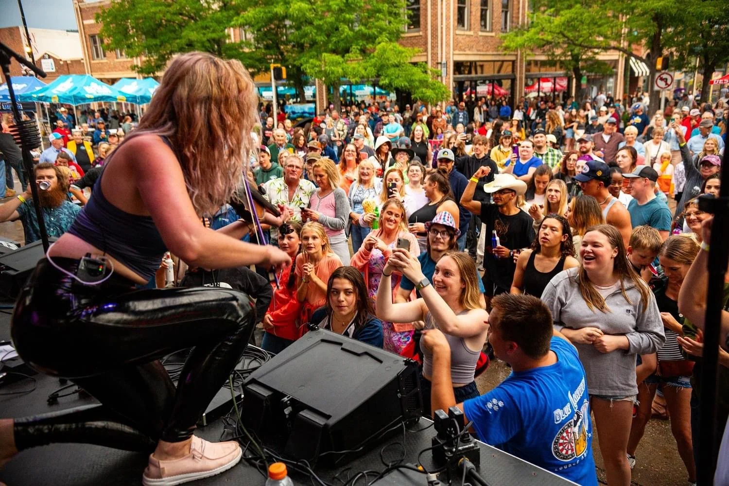 A female singer performing with a guitar on stage at an outdoor concert, with a large crowd of people watching and taking photos, set against a backdrop of trees and buildings.