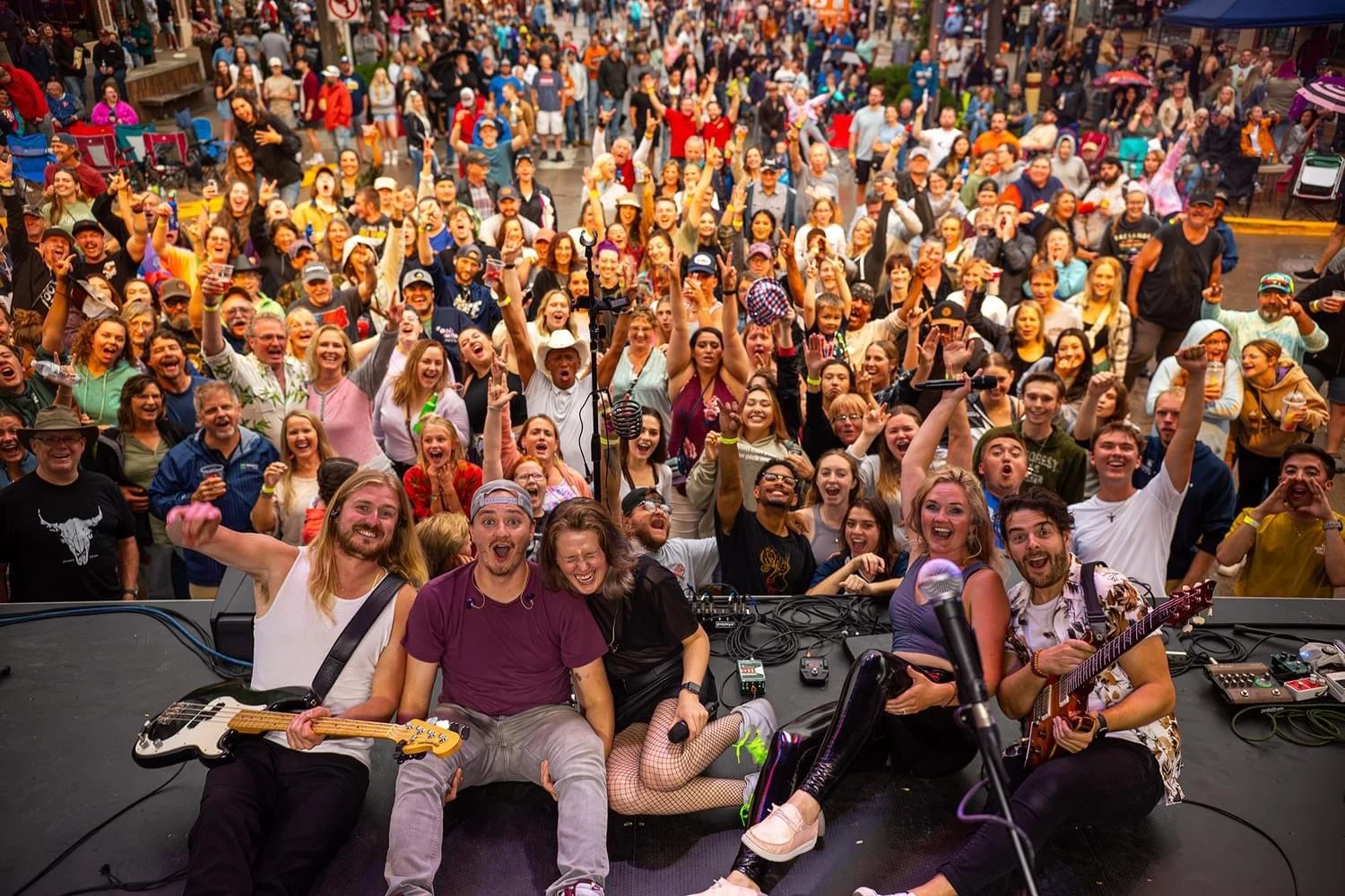 A band of musicians on stage taking a selfie with a large, cheering crowd at an outdoor concert or festival.