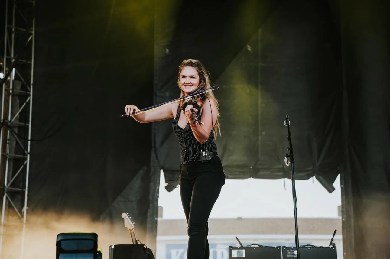 A woman playing the violin on stage at a concert, smiling, with a microphone stand and guitar in the background.
