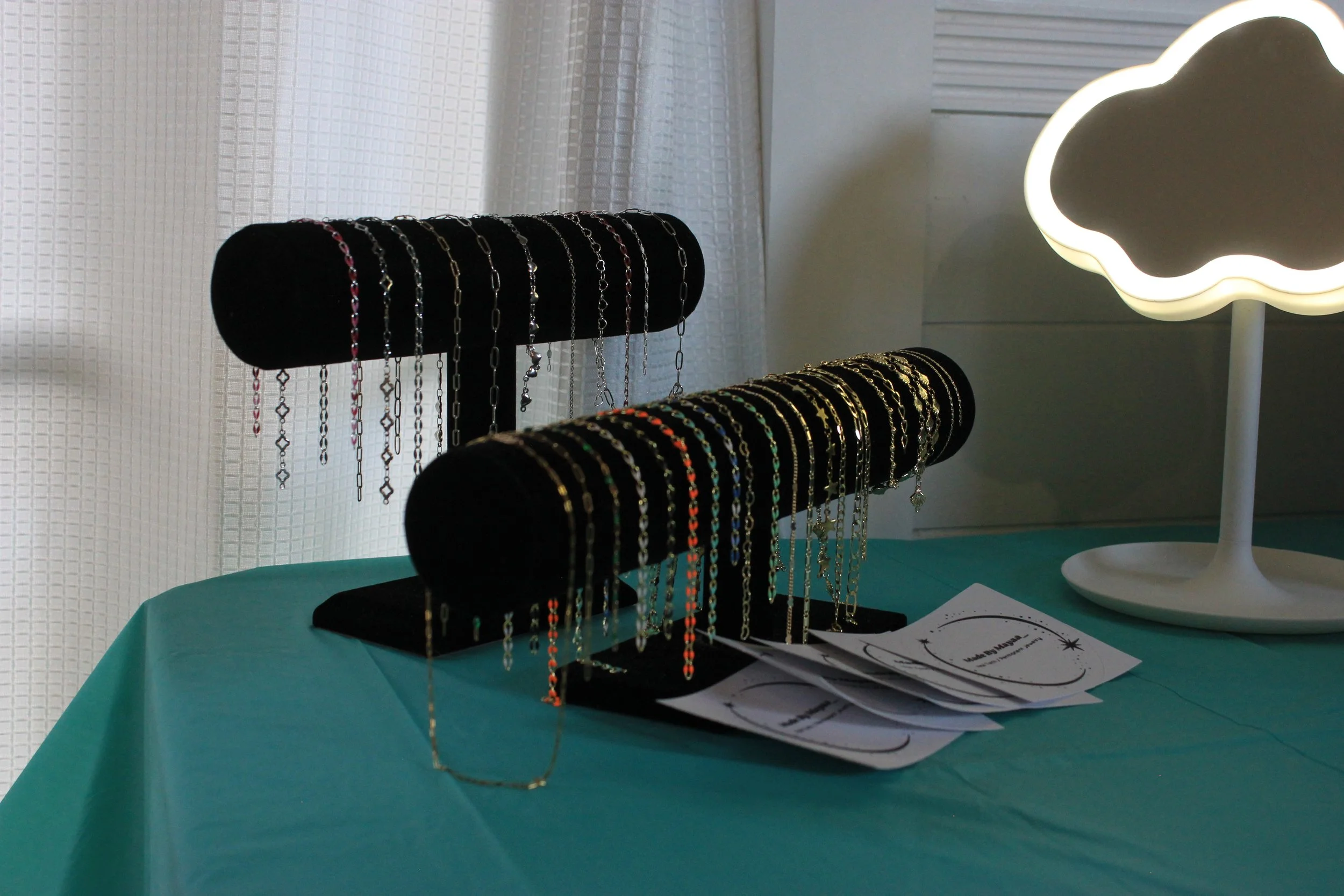 Display of assorted jewelry necklaces on black velvet busts, with a nearby white cloud-shaped lamp on a teal table.