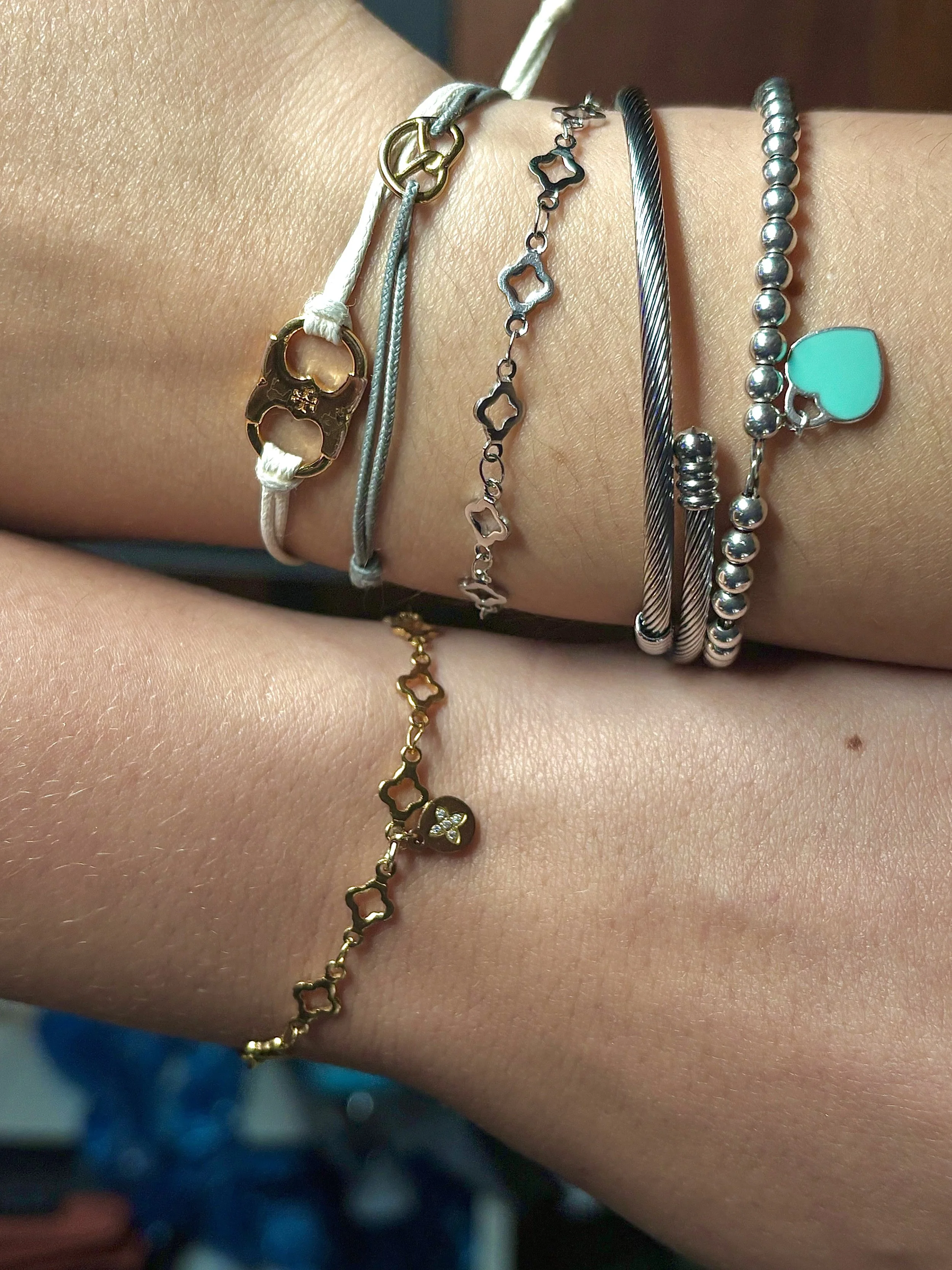 Two arms with multiple bracelets. The upper arm has five silver bracelets, including a heart-shaped turquoise bracelet. The lower arm has a gold bracelet with star-shaped links and a small star charm with tiny stones.