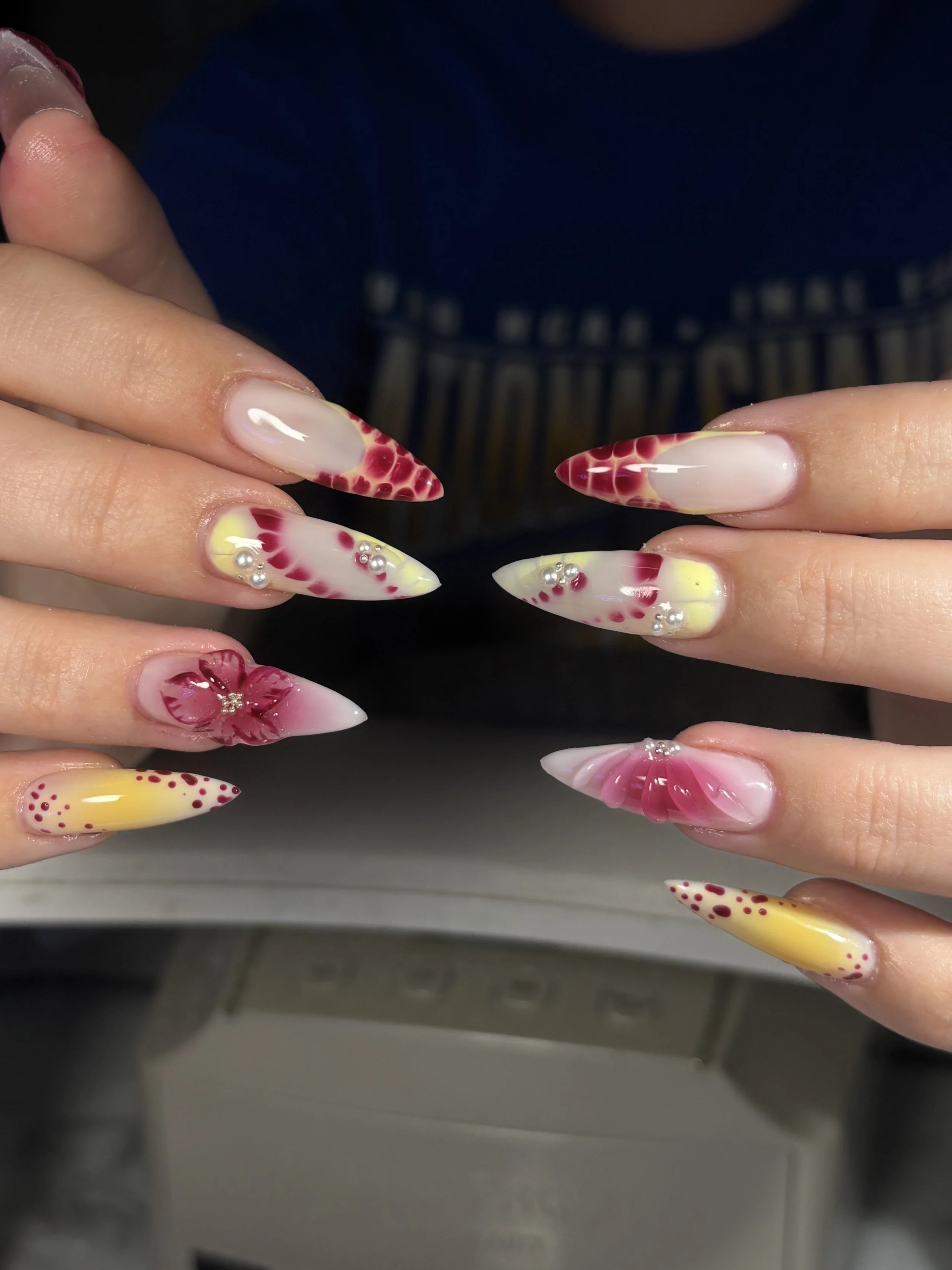 Close-up of a person's hand showing manicured nails with pink, white, and yellow nail art, including floral designs and rhinestones.