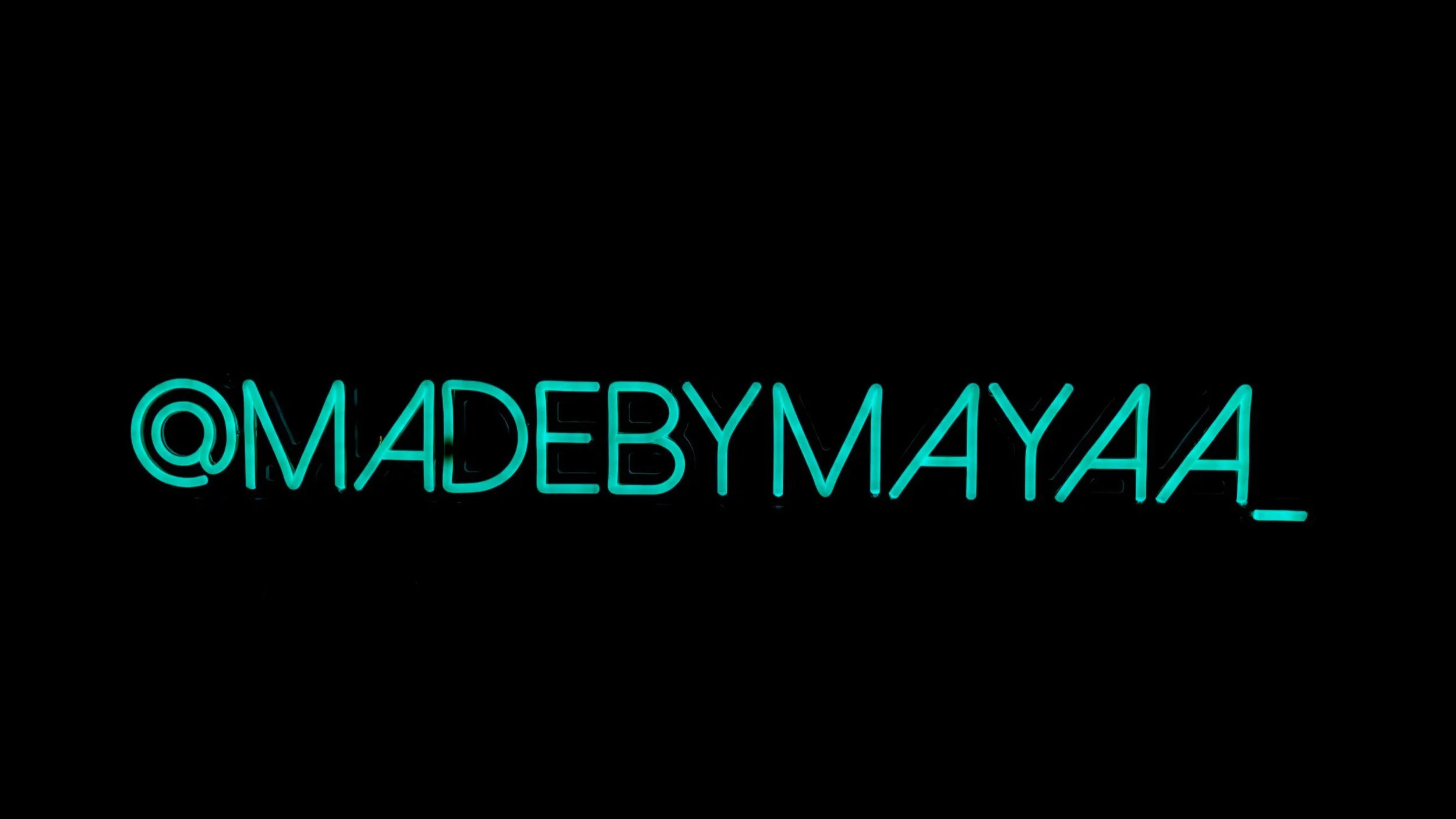 Blue neon sign displaying the social media handle '@MADEBYMAYAA_' against a black background.