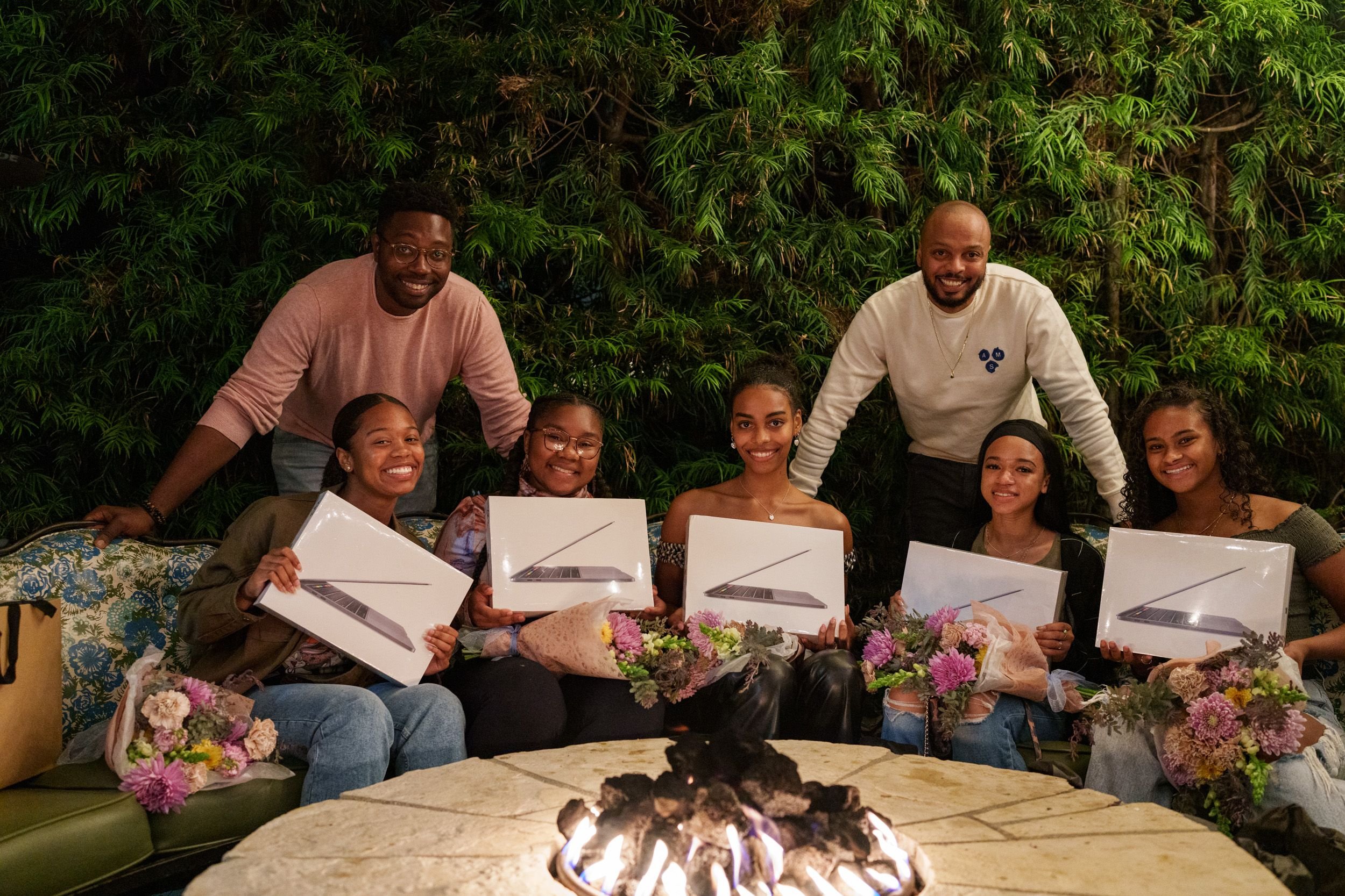 Group of five women holding gifts, accompanied by two men, sitting by a fire pit, with flowers on their laps and a green tiled wall in the background.