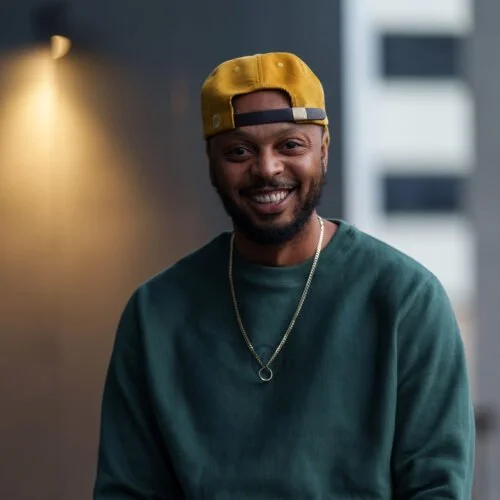 A smiling man wearing a yellow cap backwards, a green sweatshirt, and a silver chain, standing indoors.